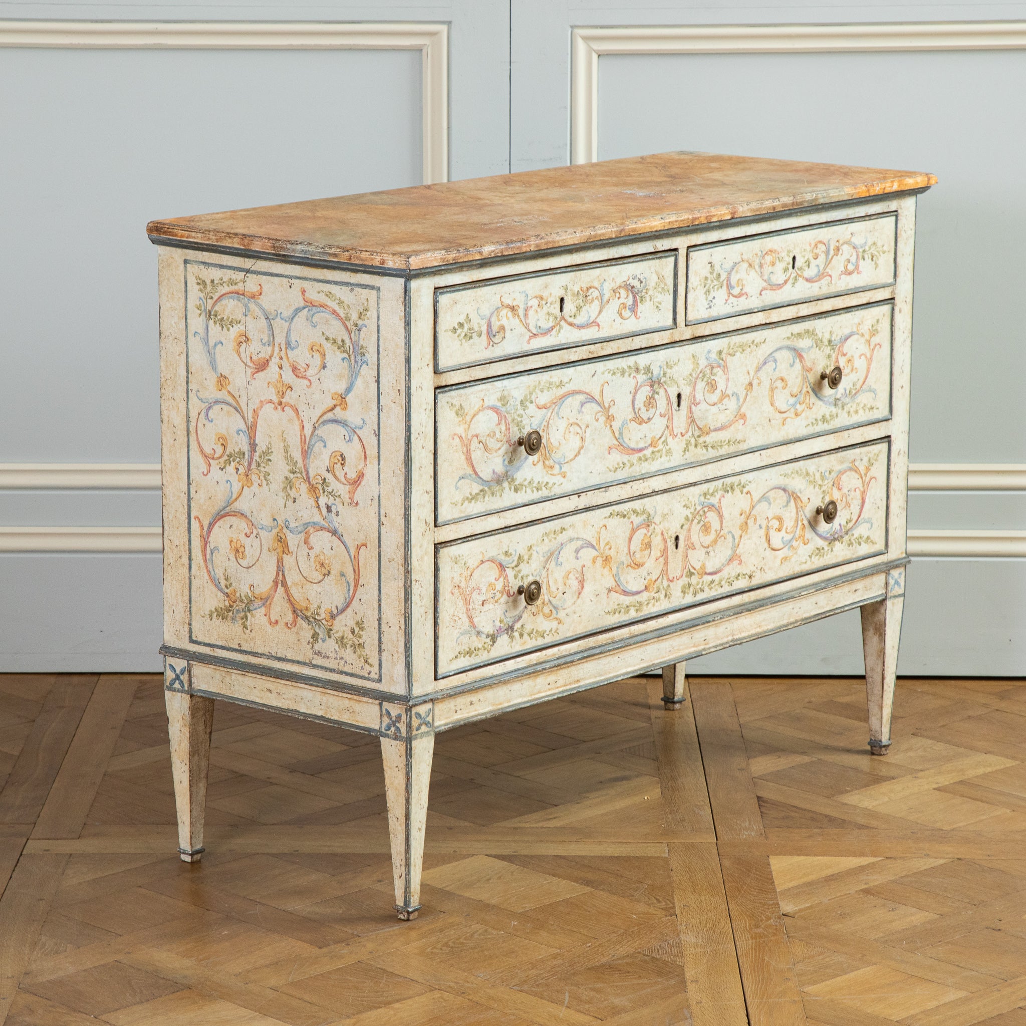 A Late 19th Century Venetian commode, hand painted in the Neoclassical style featuring fine scroll work in hues of brick pinks, yellow ochres, olive greens and blues on an old white naturally aged, craquelure background. The overall warm tones of