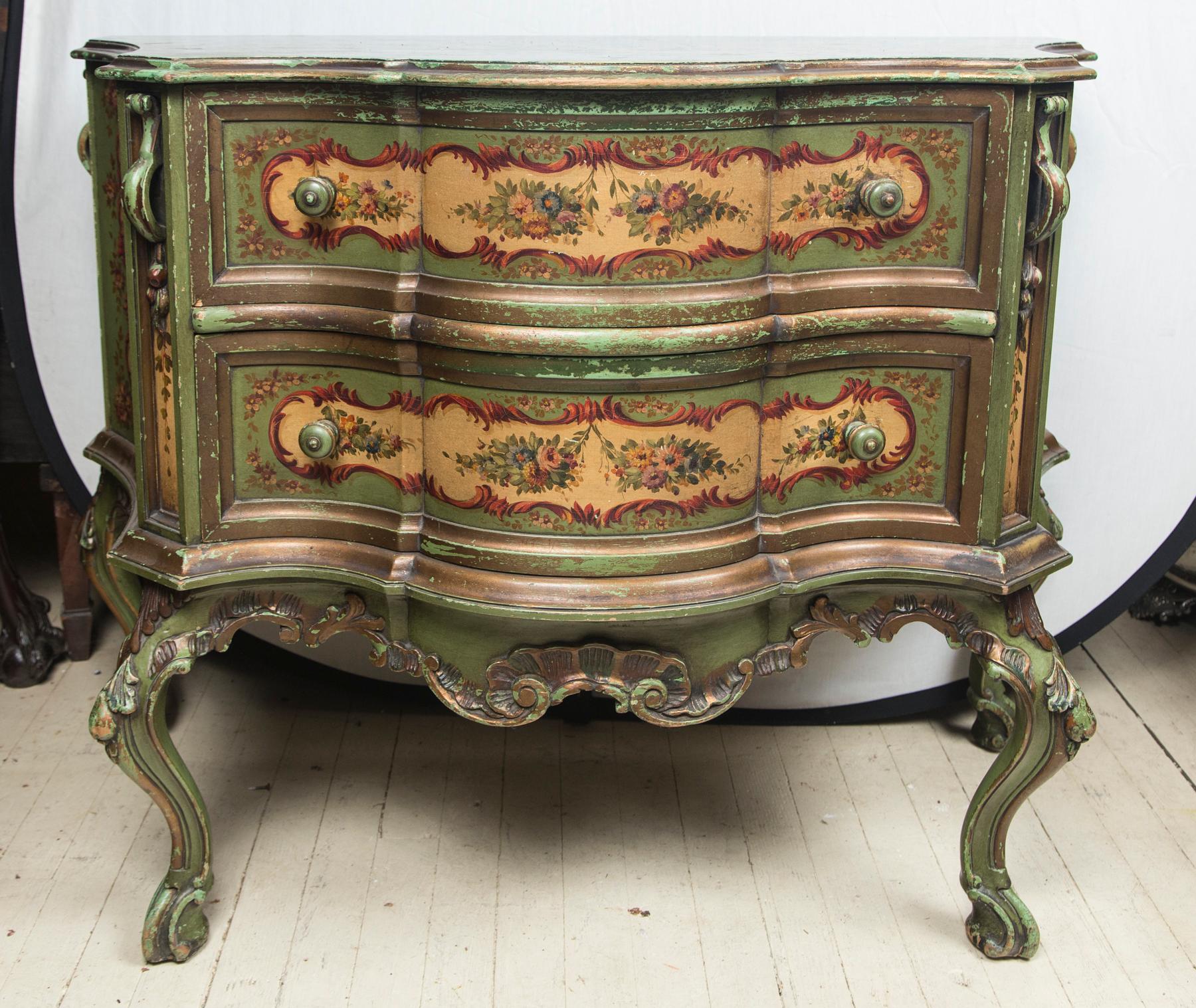 The serpentine commode with 2 drawers. The top painted faux marble. The drawers and sides painted with florals set within garland cartouches. Cabriole legs adorned with carved leaves and paint decoration. Carved ornamentation at the top of each