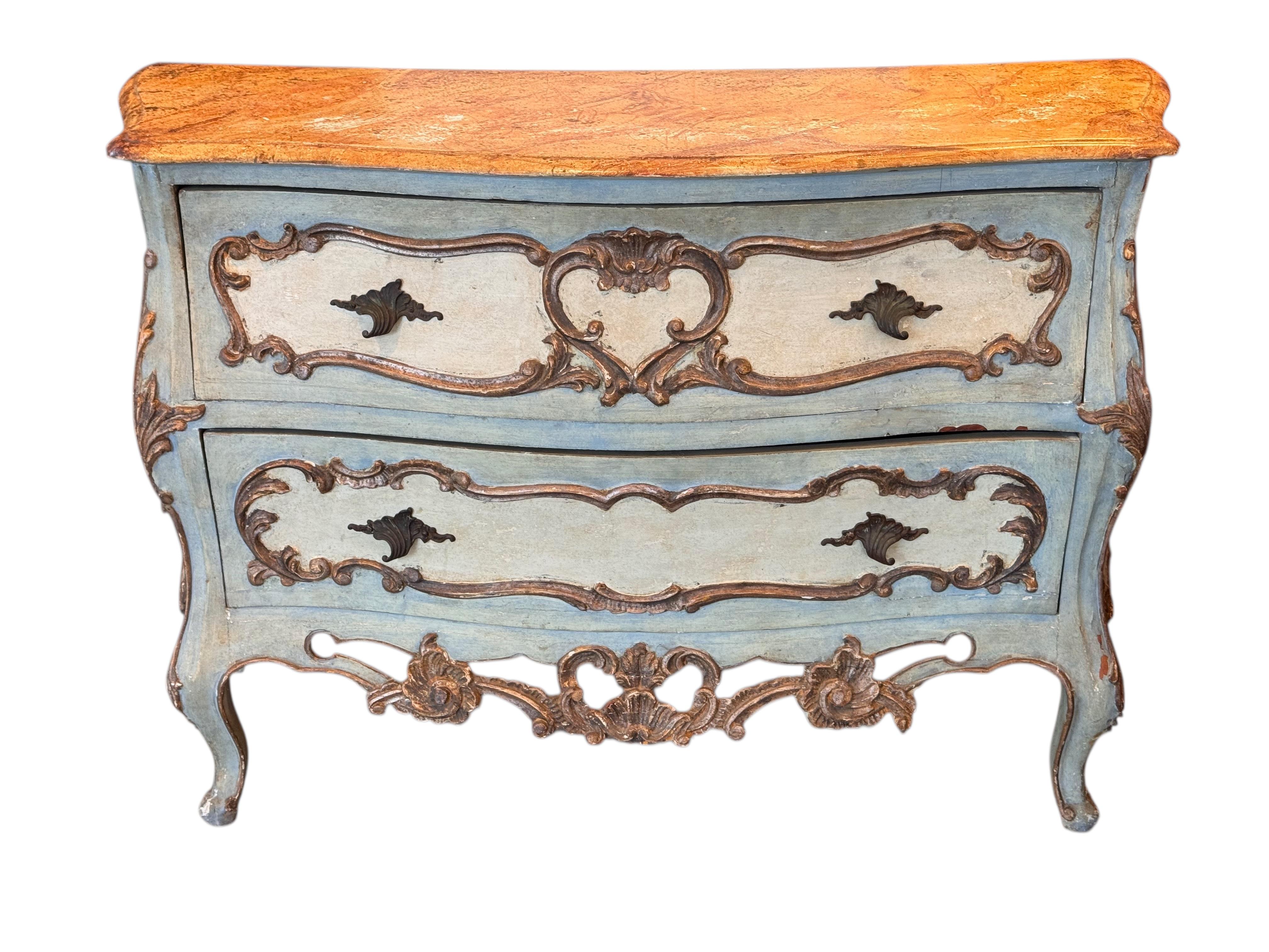 Decorative two-drawer Venetian commode, featuring a soft blue painted finish with gilt accents and elaborate carved floral and scrollwork motifs. The shaped drawers and cabriole legs emphasize its baroque influence, while the painted surface adds a
