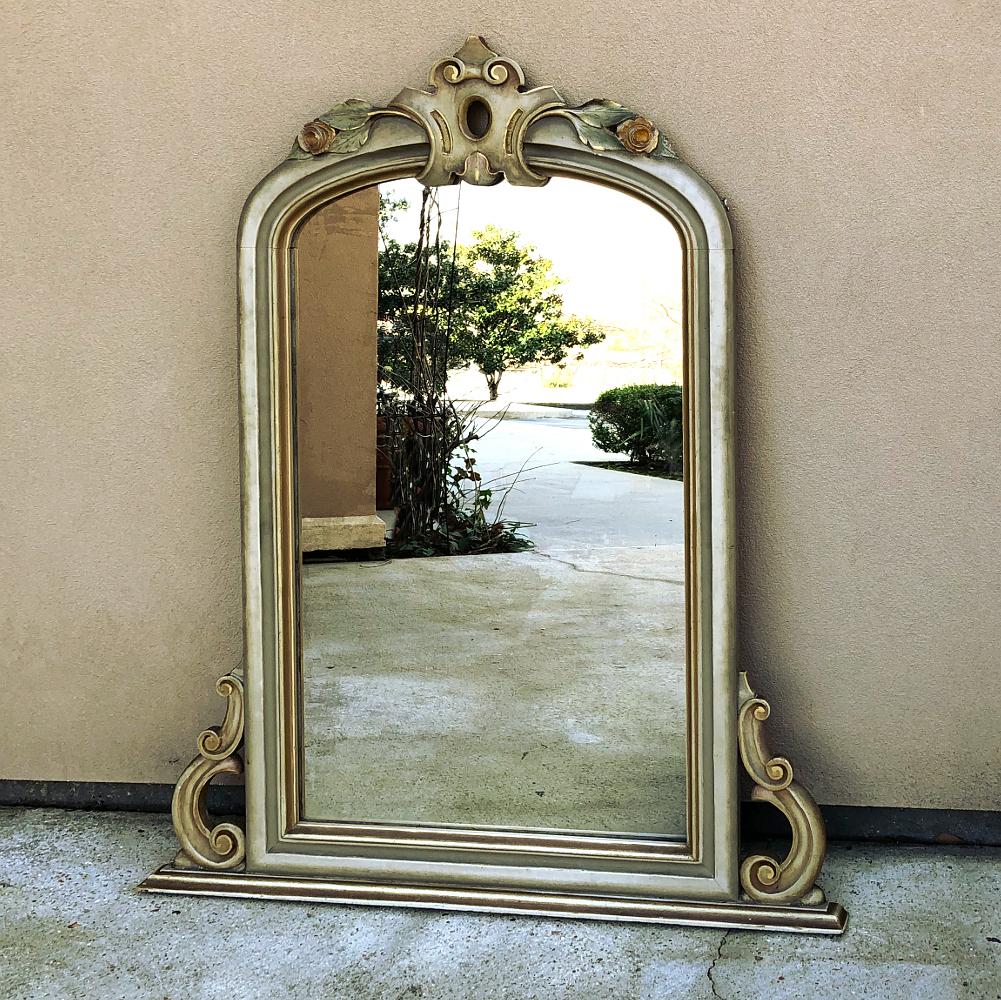 19th century Venetian painted mirror features a bold heraldic crest atop the gracefully arched crown, flanked by cascading floral and foliate sprays accented with blushes of color. The overall taupe and light gray color of the framework is