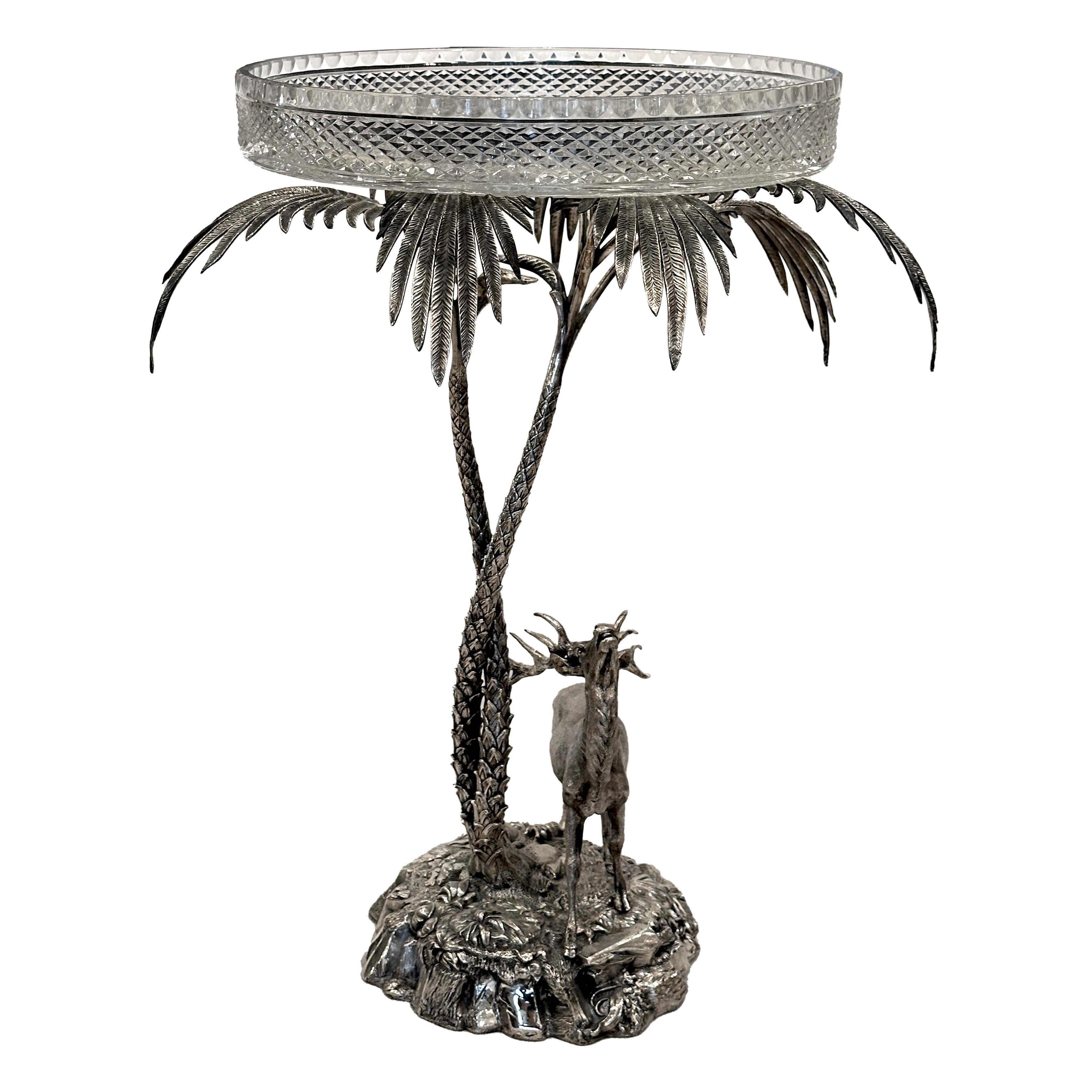Questo pezzo in bronzo argentato finemente fuso raffigura un maestoso cervo in piedi su una base rocciosa, che osserva due grandi palme intrecciate, sormontate da un vassoio di cristallo. Sul lato è presente il timbro dell'argentiere Elkington & Co.