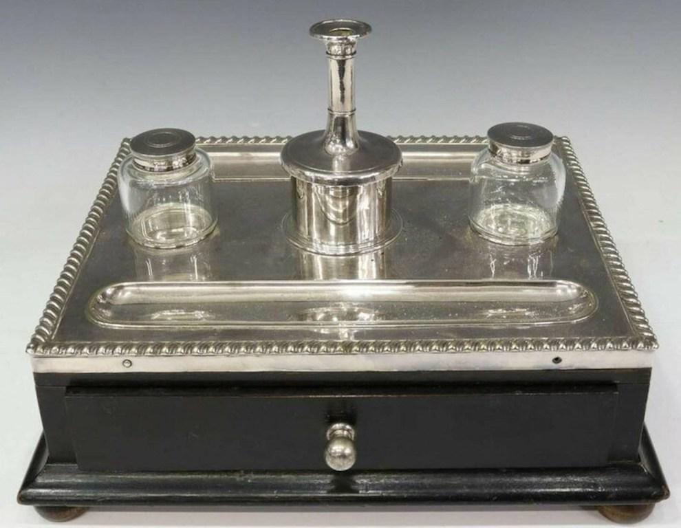 A charming Victorian era English silver plate desk stand, born in the late 19th century, having two glass inkwell pots, candlestick, recessed pen wells, gadrooned border, ebonized case fitted with a single drawer, rising on bun feet.

Clean,