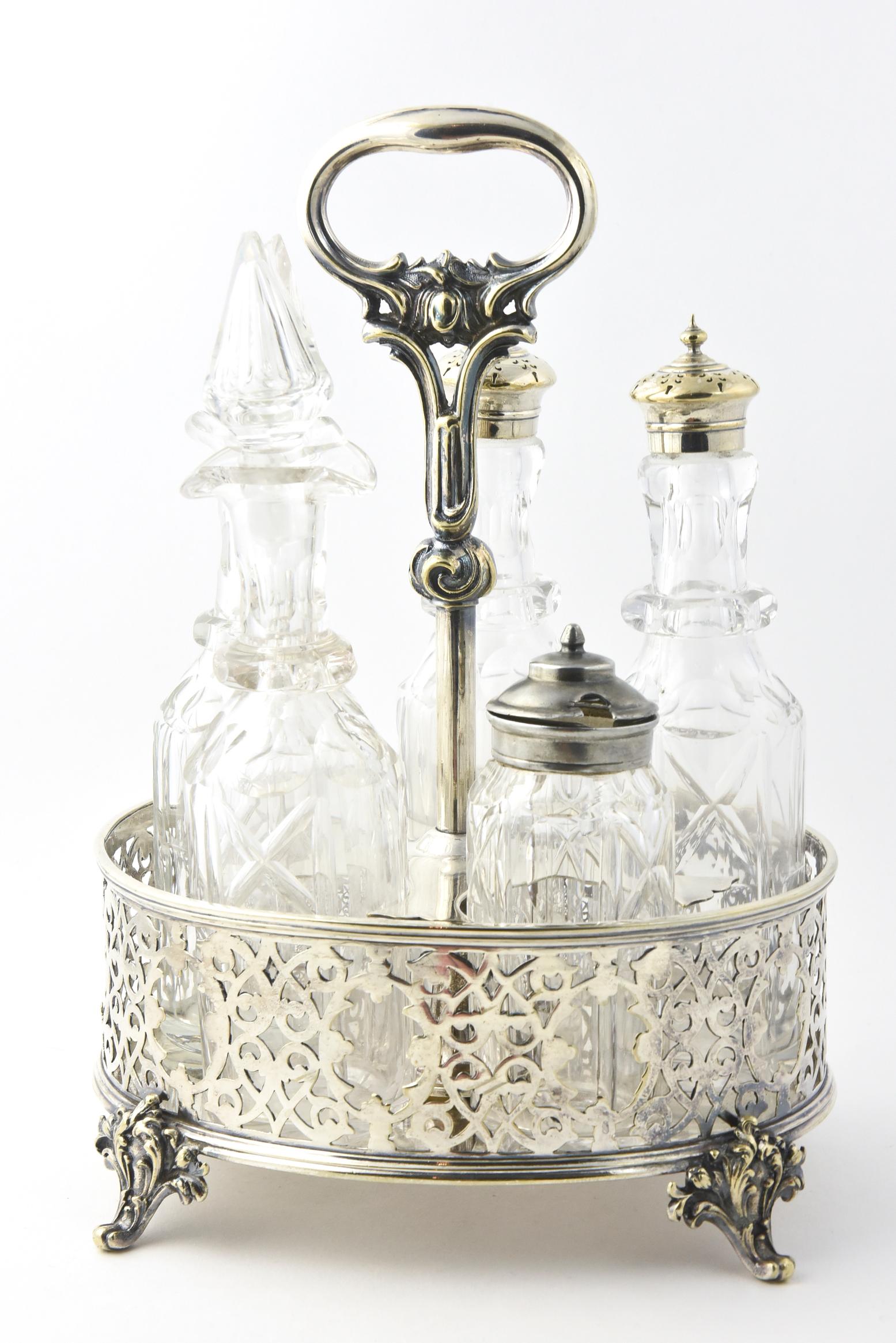 Antique silver plate caster set with five cut-crystal bottles: salt, pepper, oil, vinegar, and mustard. The bottles are displayed in a fitted silver plate stand with a central handle. White film on one glass; one glass stopper is chipped.