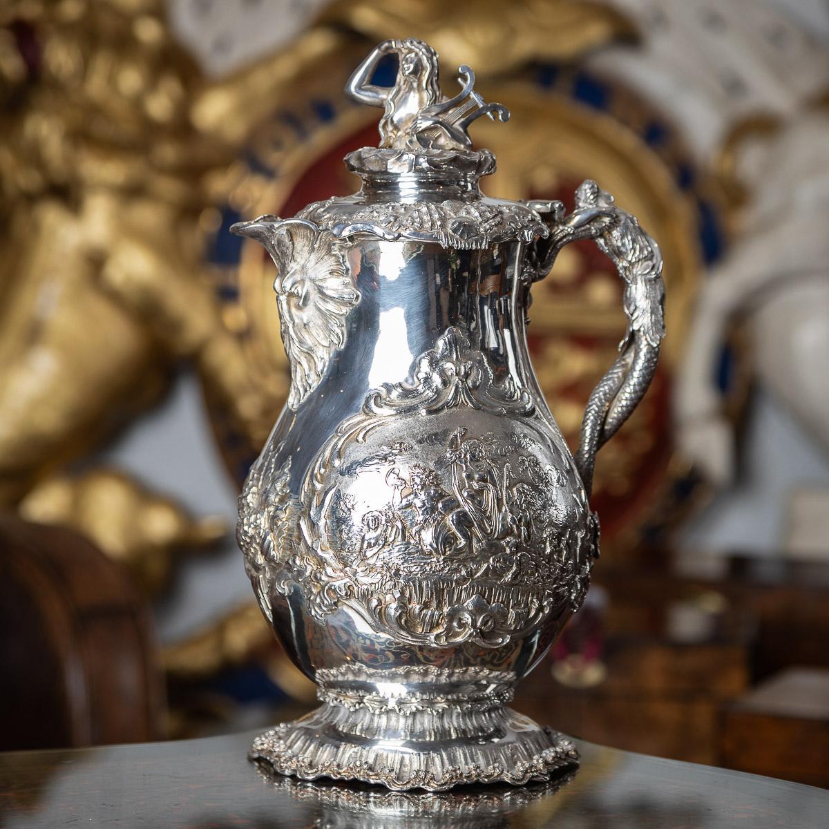 Antique mid-19th Century Victorian impressively large solid silver water jug, the generously adorned body features intricate relief depicting nautical scenes, elaborate scrolls, and mythical dolphins. Standing proudly on a spreading foot embellished