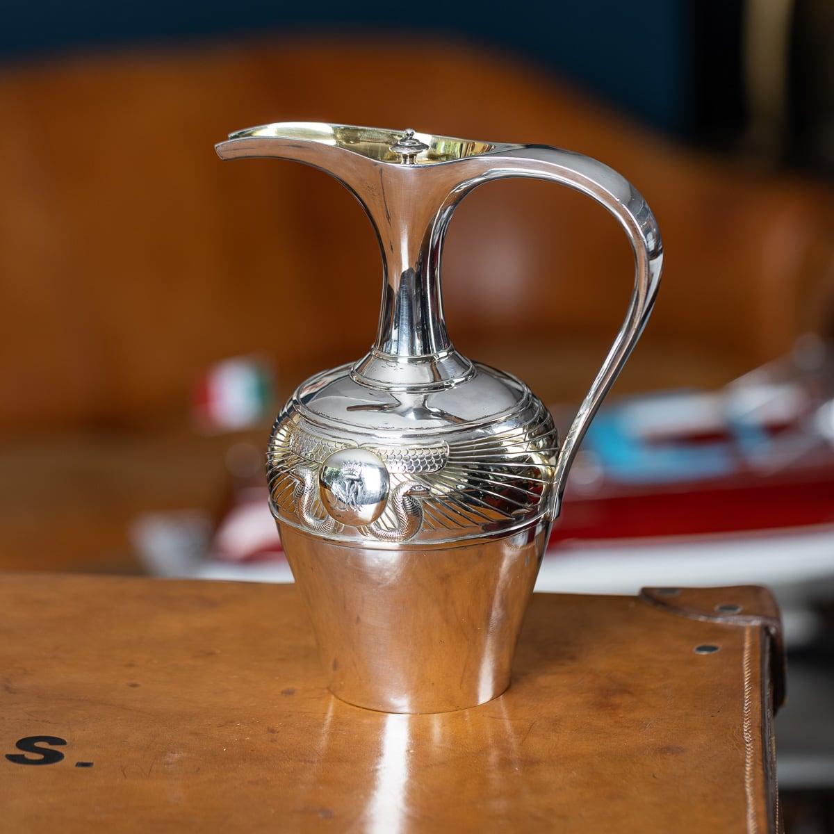 Antigua jarra de vino victoriana del siglo XIX, de plata maciza dorada, de estilo egipcio, con un par de serpientes en el centro flanqueadas por alas, cartela grabada con las iniciales coronadas. La parte superior presenta un pico alargado con una