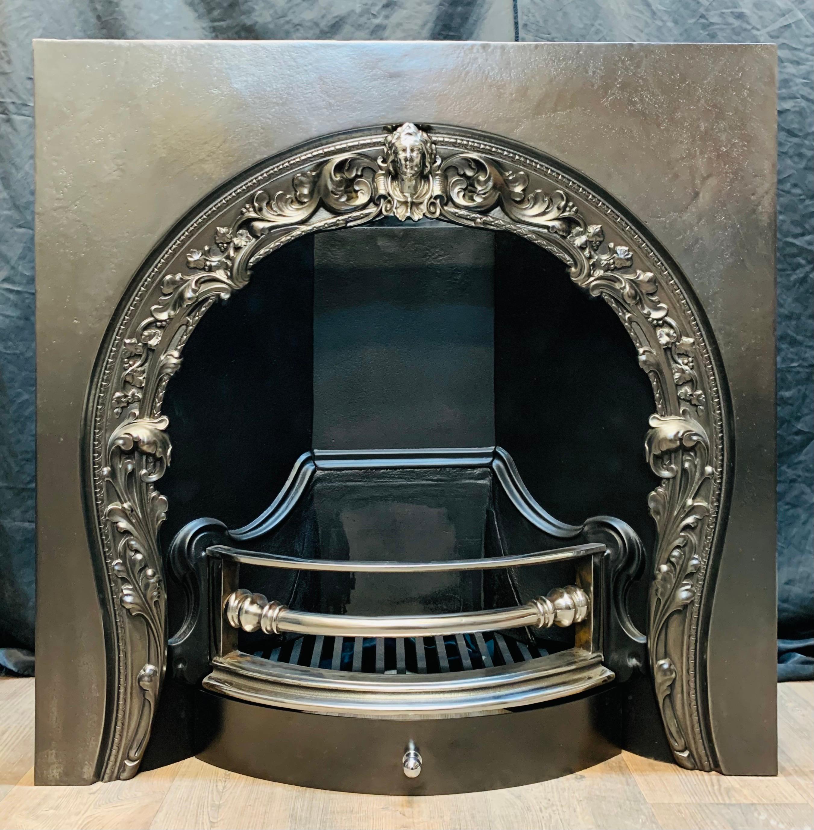 A large 19th century Victorian style arched cast iron fireplace insert. A generous outer frame with the horseshoe shaped opening displaying various leafs with a polished cast female mask to the center arch, the curved and polished three barred grate