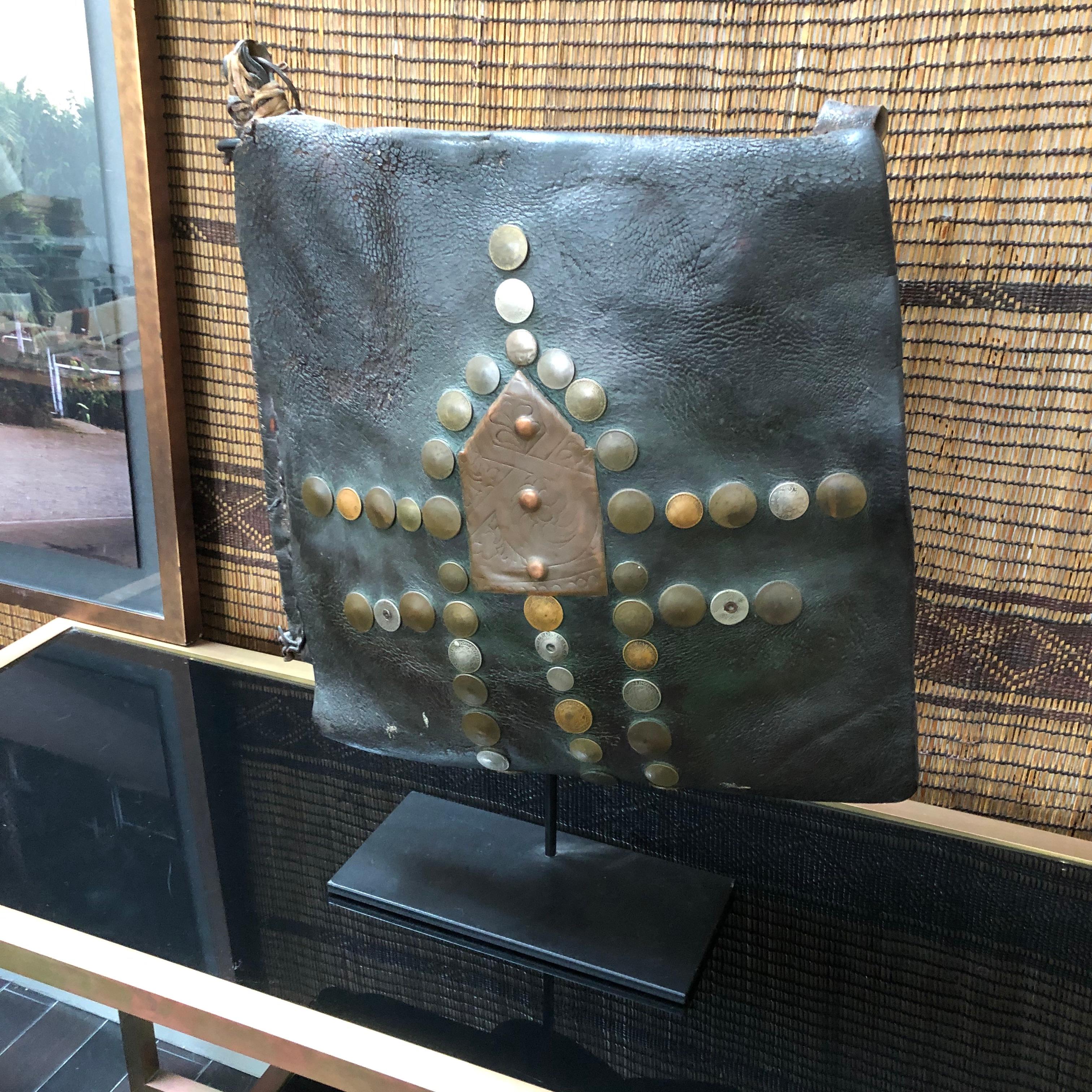 A vintage handcrafted leather water carrier bag from Morocco on a stand. Handmade with camel hide and decorated with antiqued coins from around the world. 

Traditionally made for water sellers in order to carry water through markets and sell to