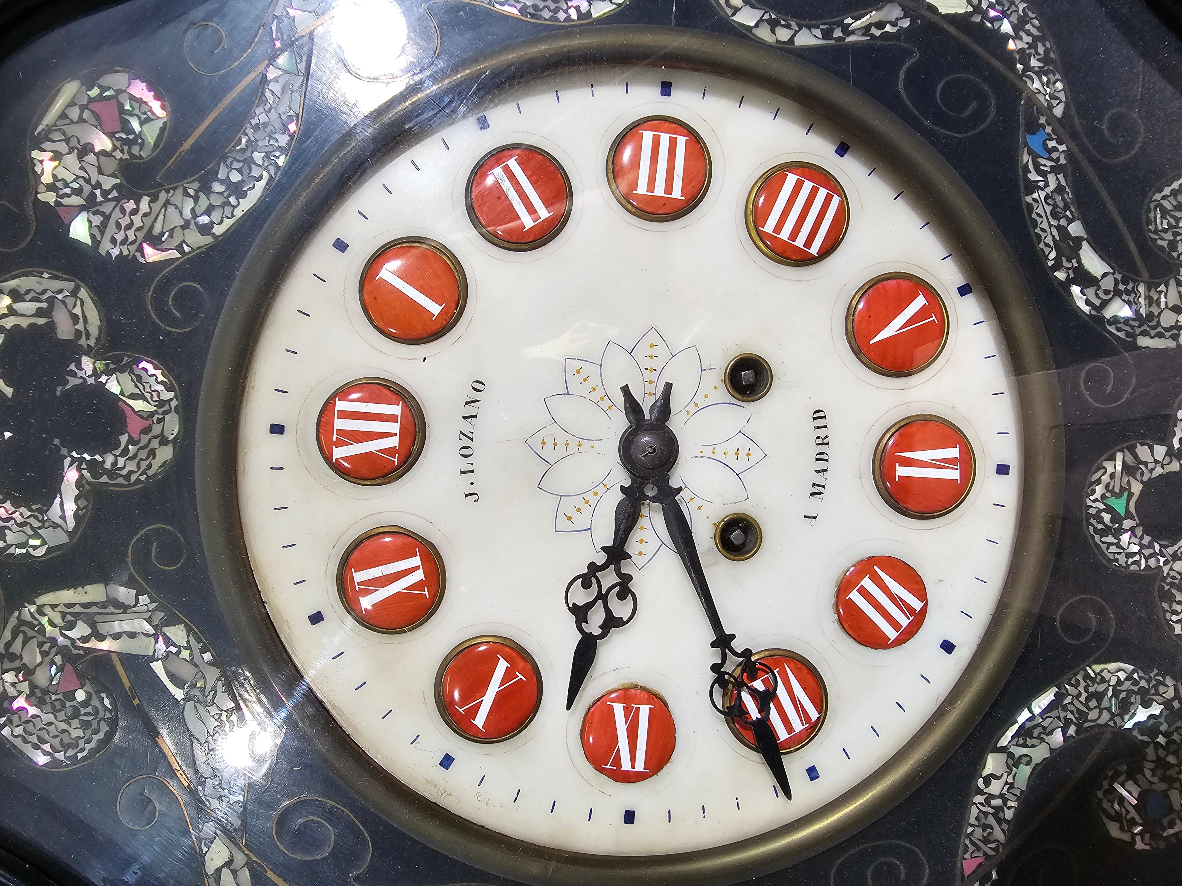 iese elegante Wanduhr aus dem 19. Jahrhundert, signiert von J. Lozano in Madrid, ist ein beeindruckendes Beispiel für spanische Handwerkskunst. Aus wunderschön ebonisiertem Holz gefertigt und mit exquisiten Perlmutt-Intarsien verziert, verbindet er