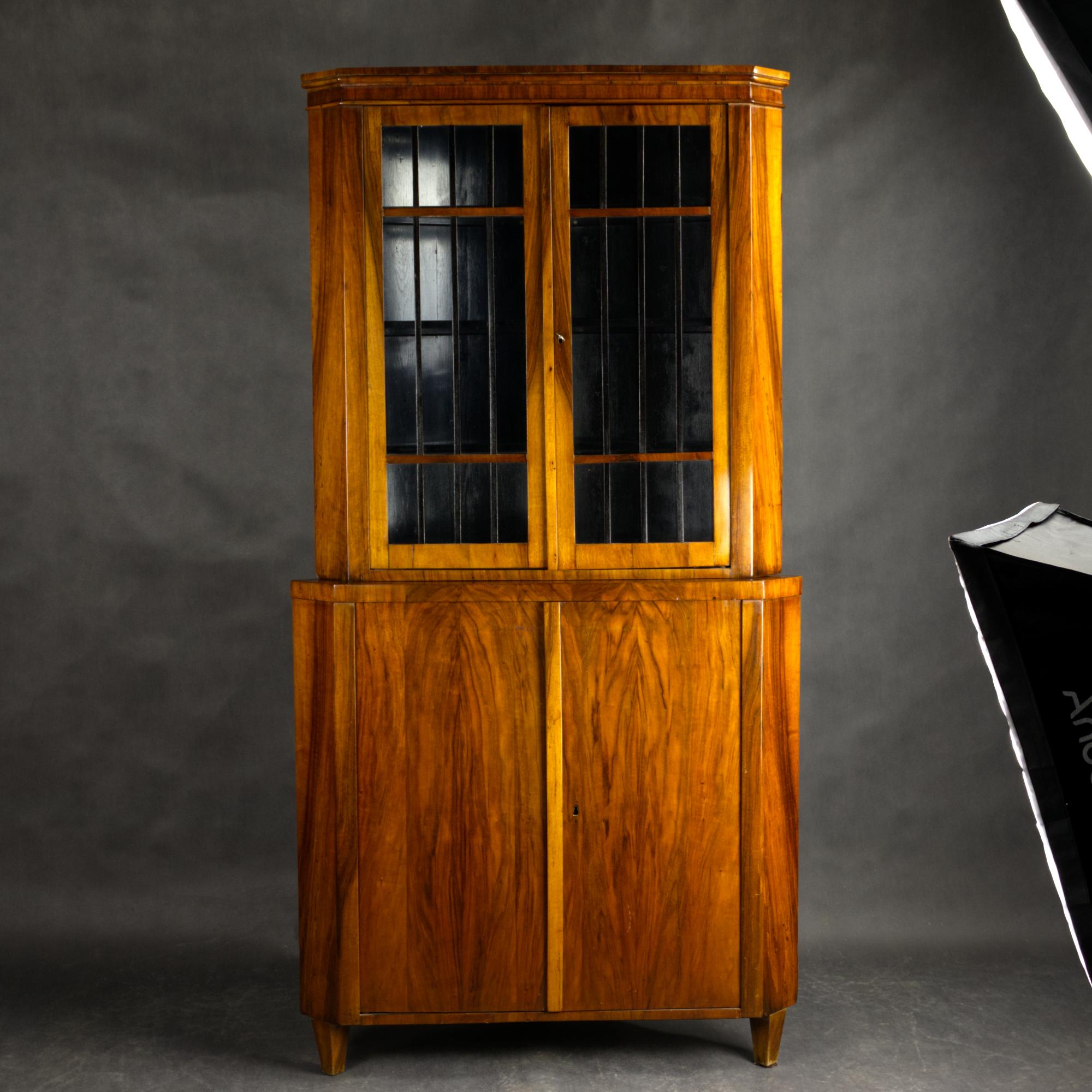 Eine auffällige Eckvitrine aus dem späten Biedermeier, gefertigt aus schön gemasertem Nussbaum mit einer warmen, natürlichen Patina. Im oberen Teil befinden sich zwei Glastüren mit eleganten vertikalen Sprossen, die sich ideal zur Präsentation von