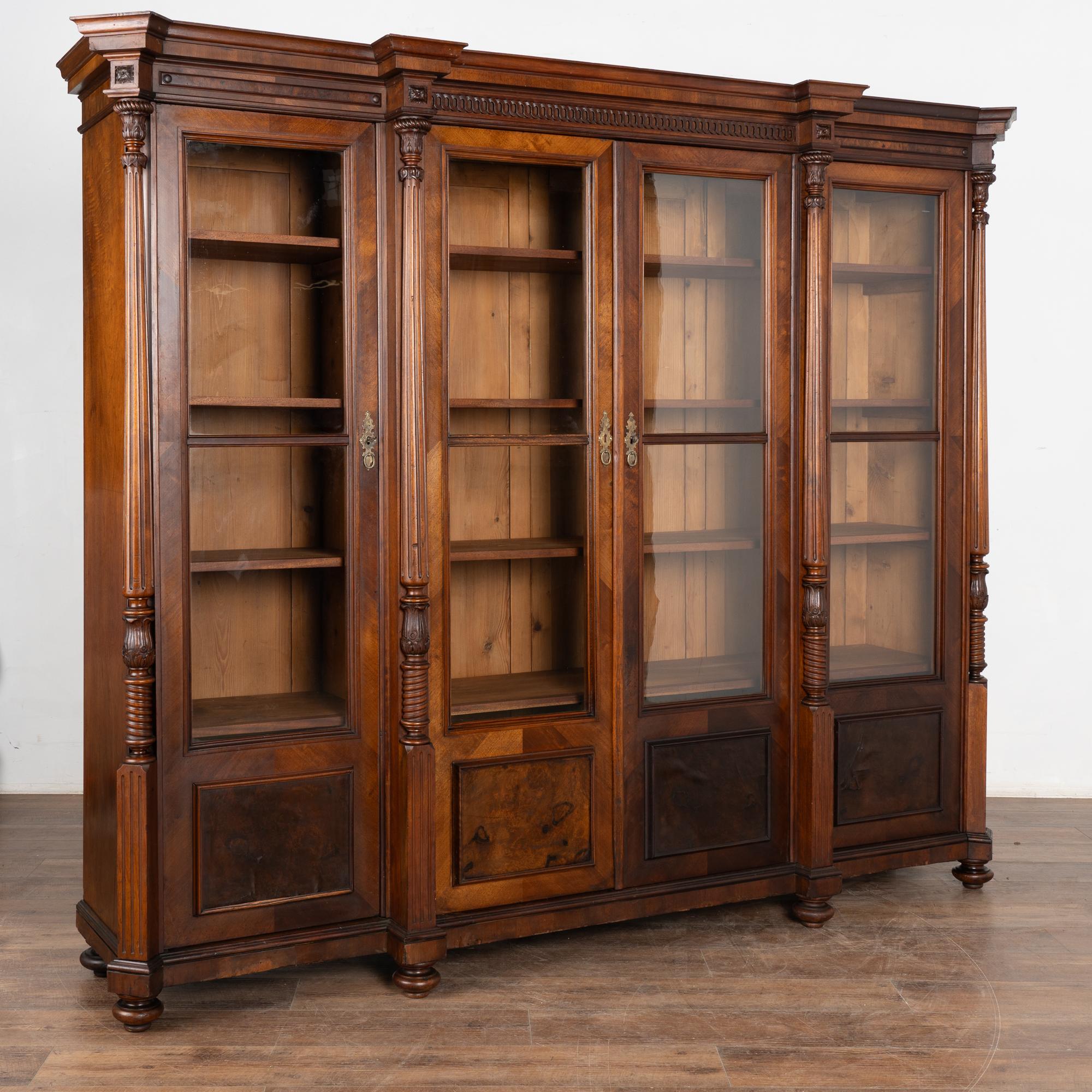 Les quatre longues portes vitrées sont complétées par des panneaux de placage inférieurs dans cette grande bibliothèque en noyer. Flanquée de colonnes tournées et d'une corniche décorative, cette vitrine traditionnelle présente une ligne statuaire