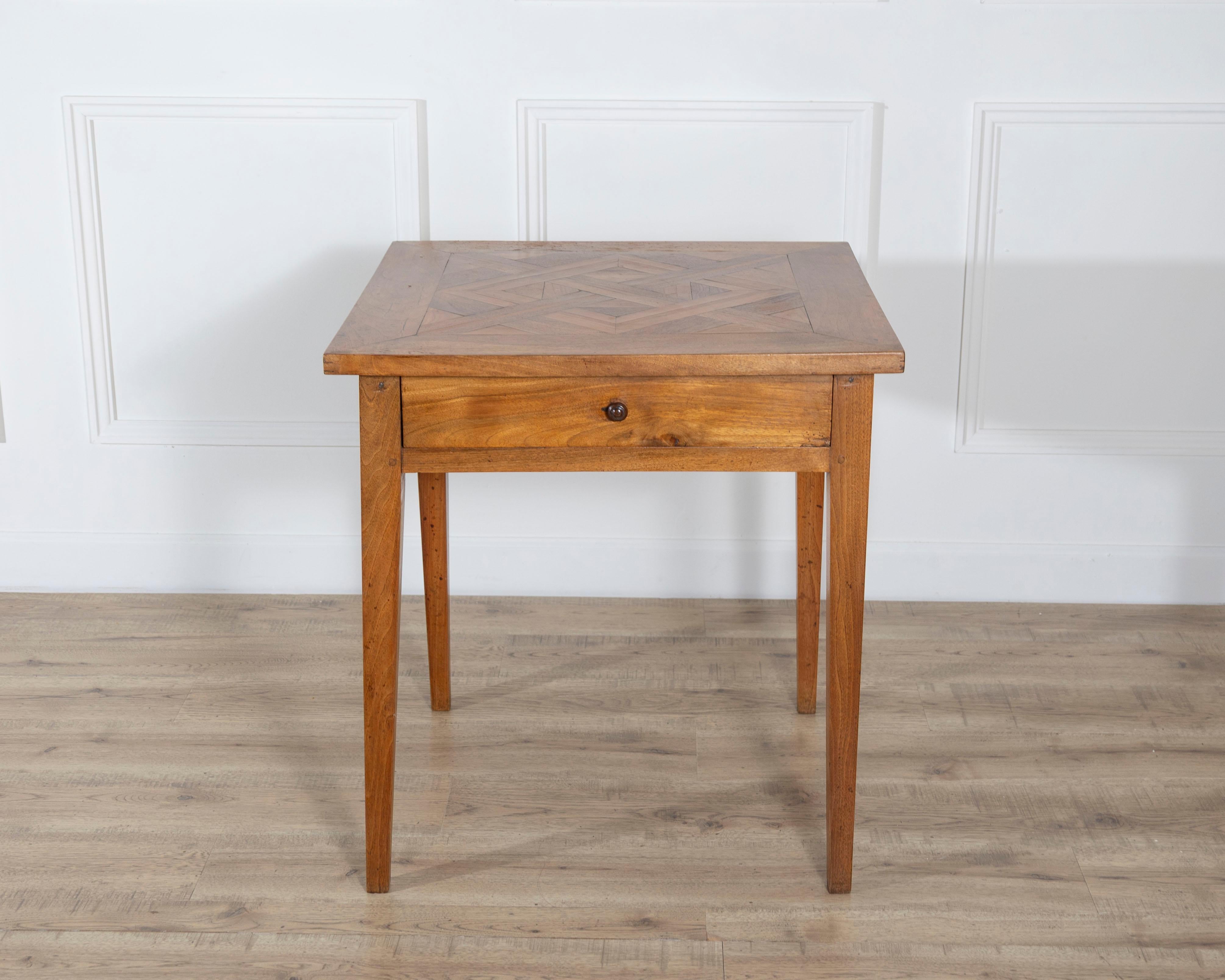 This square walnut table features a geometric inlaid top composed of bookmatched and angled walnut veneers that create a clear, symmetrical pattern. The frame is constructed in solid walnut with a warm, even finish. A single drawer is integrated