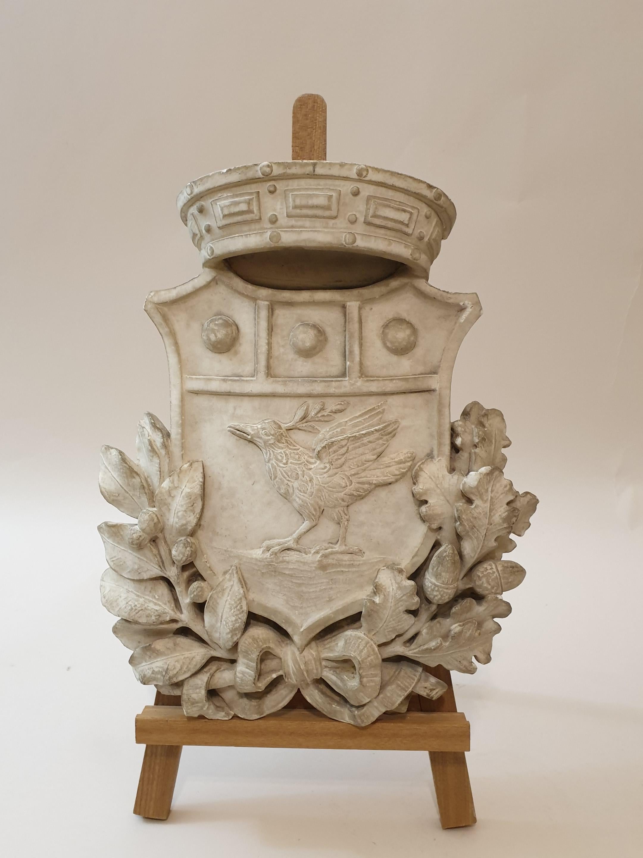 A heraldic coat of arms in white marble finely carved in bas-relief depicting a dove with an olive tree, inside a shield with crown over it and lower leaves of laurel and oak.
