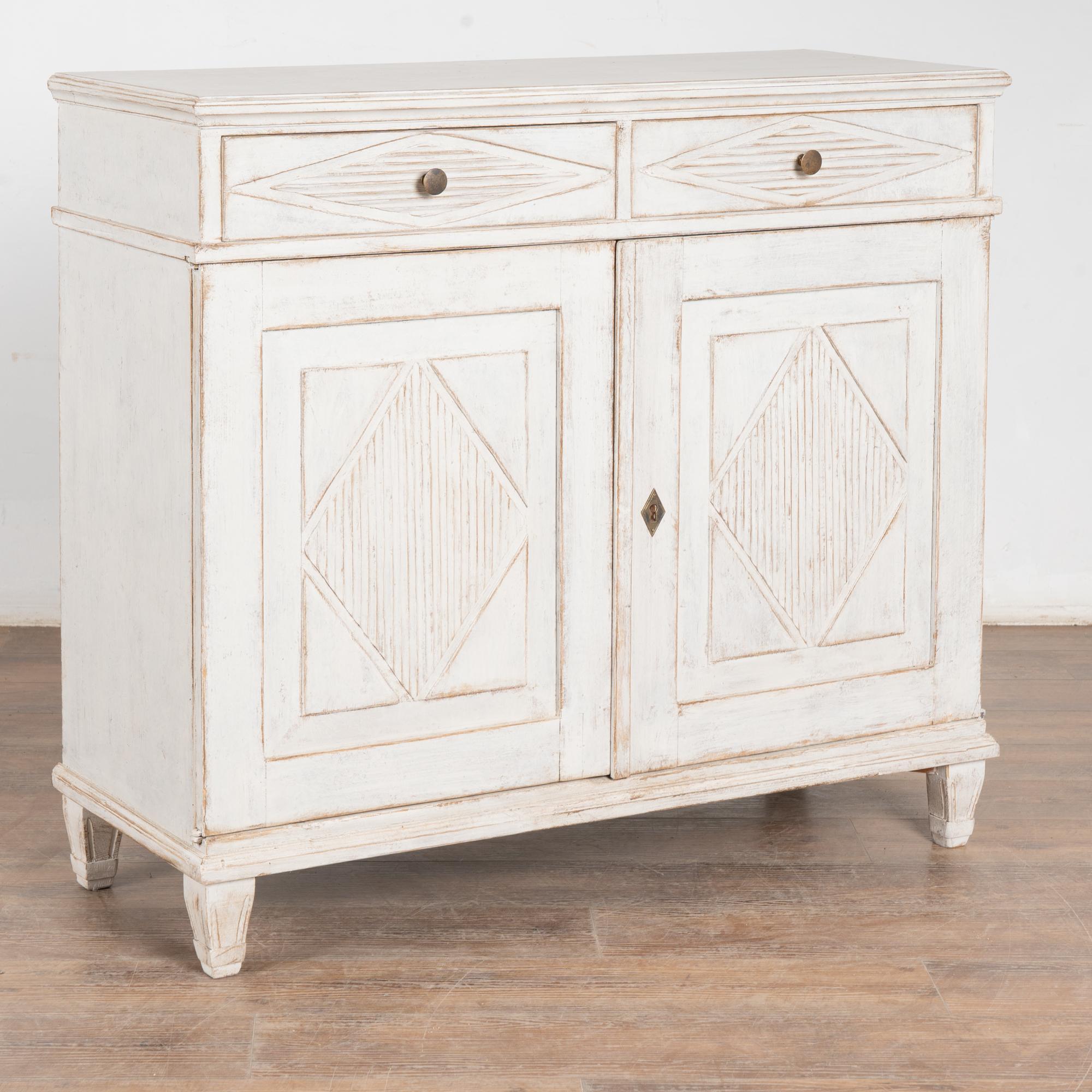 Gustavian pine sideboard or buffet with traditional decorative fluted diamond panels on cabinet doors. 
Later professionally painted in layered shades of antique white and lightly distressed to fit the age and grace of this Swedish sideboard.
Brass