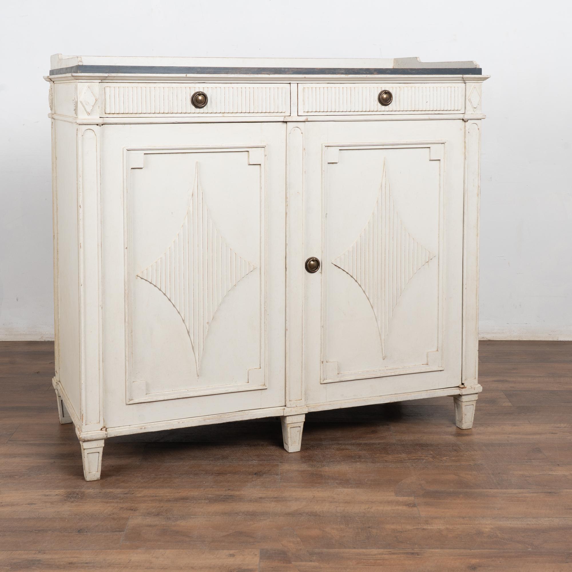 Gustavian pine sideboard or buffet with traditional decorative fluted diamond panels on cabinet doors raised on tapered feet. 
Later professionally painted in layered shades of antique white (soft gray undertones), and lightly distressed to fit the