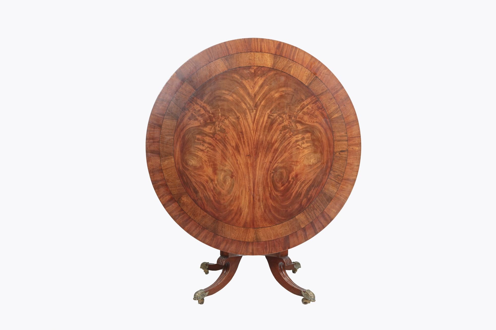 19th Century William IV mahogany circular tip-up centre table with flame mahogany center crossbanded with rosewood, with platform base held on four scroll and reeded splayed legs terminating on brass castors.