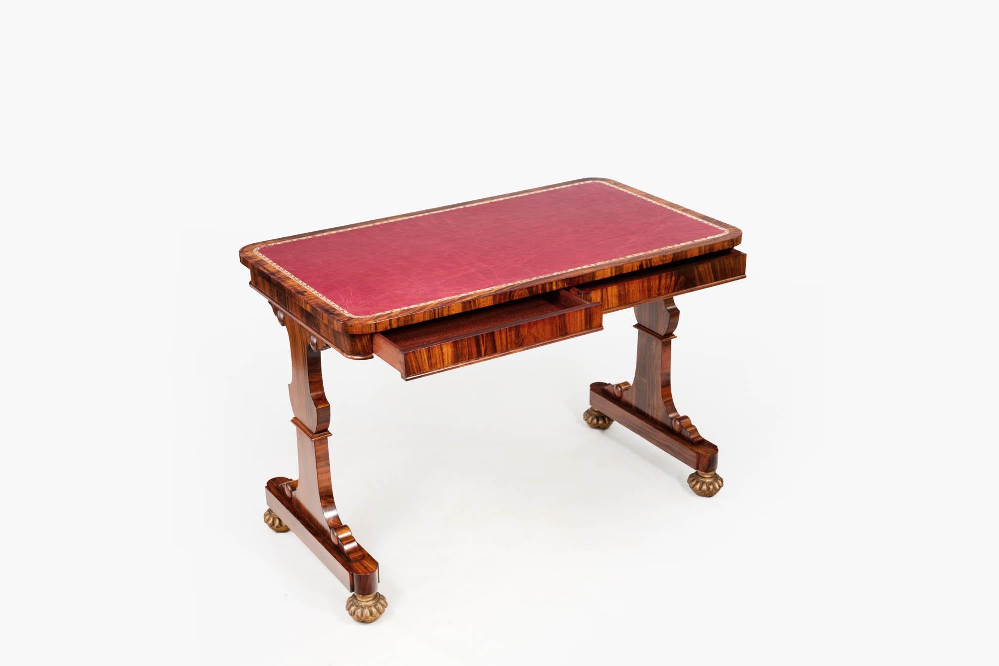19th century William IV rosewood library table. The rectangular top with rounded corners features a red tooled inset leather writing surface and two disguised drawers. Supported on twin carved uprights, this piece terminates on castors that have