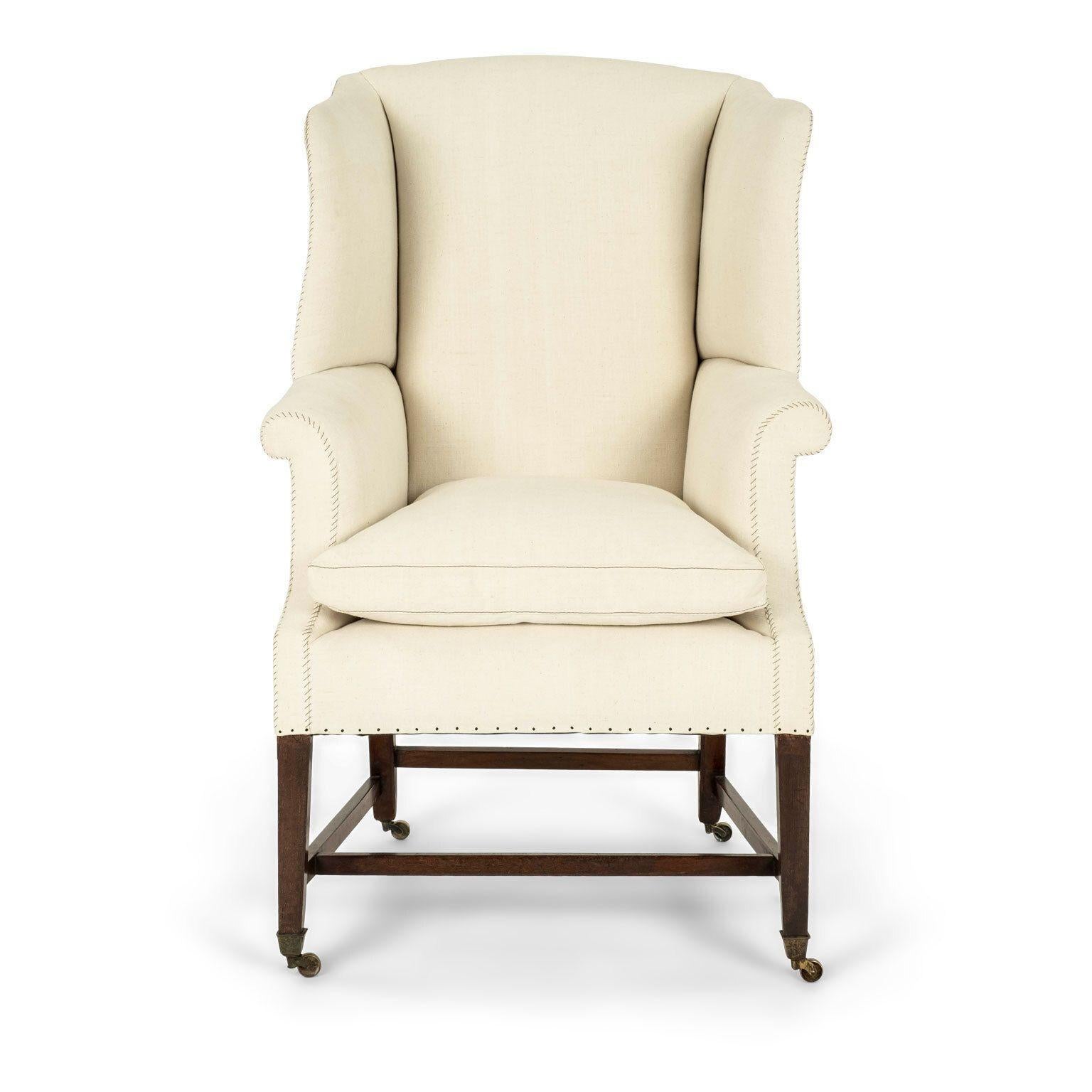George III style wingback upholstered in antique off-white linen. English porter style Georgian wingback dating to the mid-to-early 19th century. Generously proportioned scale. Rests upon squared mahogany stretchers and legs, tapered at foot in