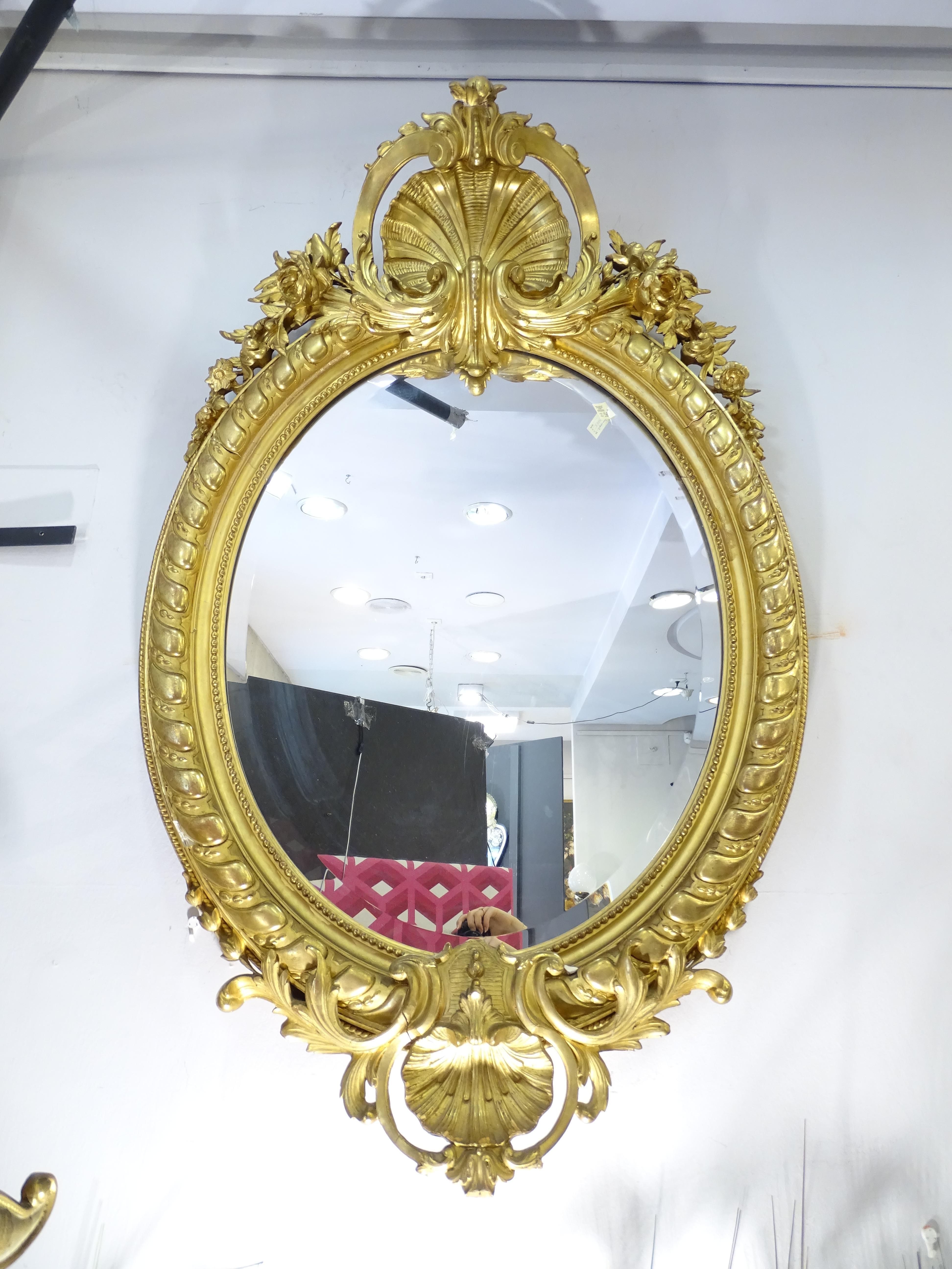 Outstanding mirror in carved and gilded wood with fine gold burnished in water with agate stone. It has an elegant oval profile made up of a double frame, the pearly interior and the chevroned exterior. The main ornament is the two central scallop