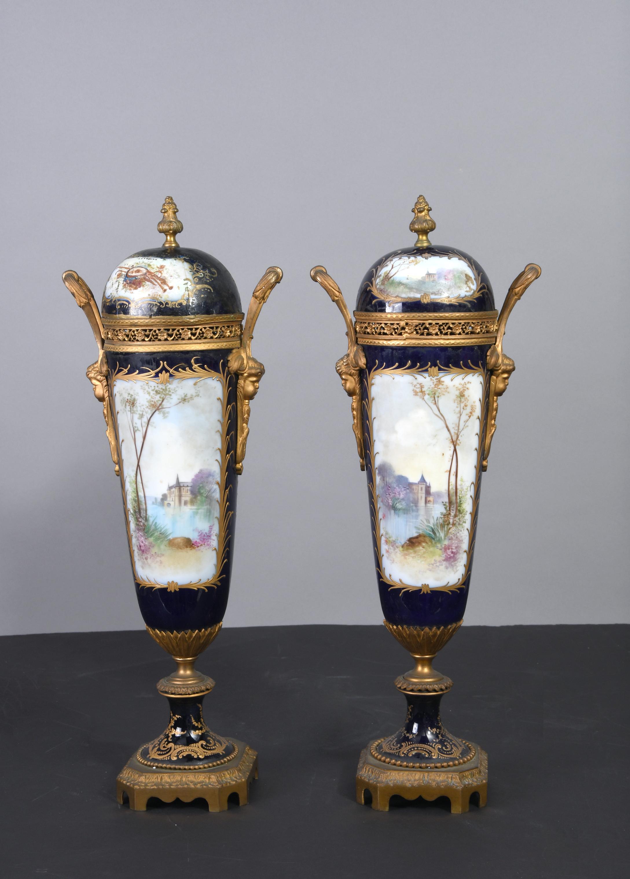 The dark blue background of the porcelain vases stand on a gilded carved octagonal pedestal. The base is decorated with gilt scrolls of acanthus leaves and foliage. The front center of the cone shaped vase is hand painted is a castle lake scene