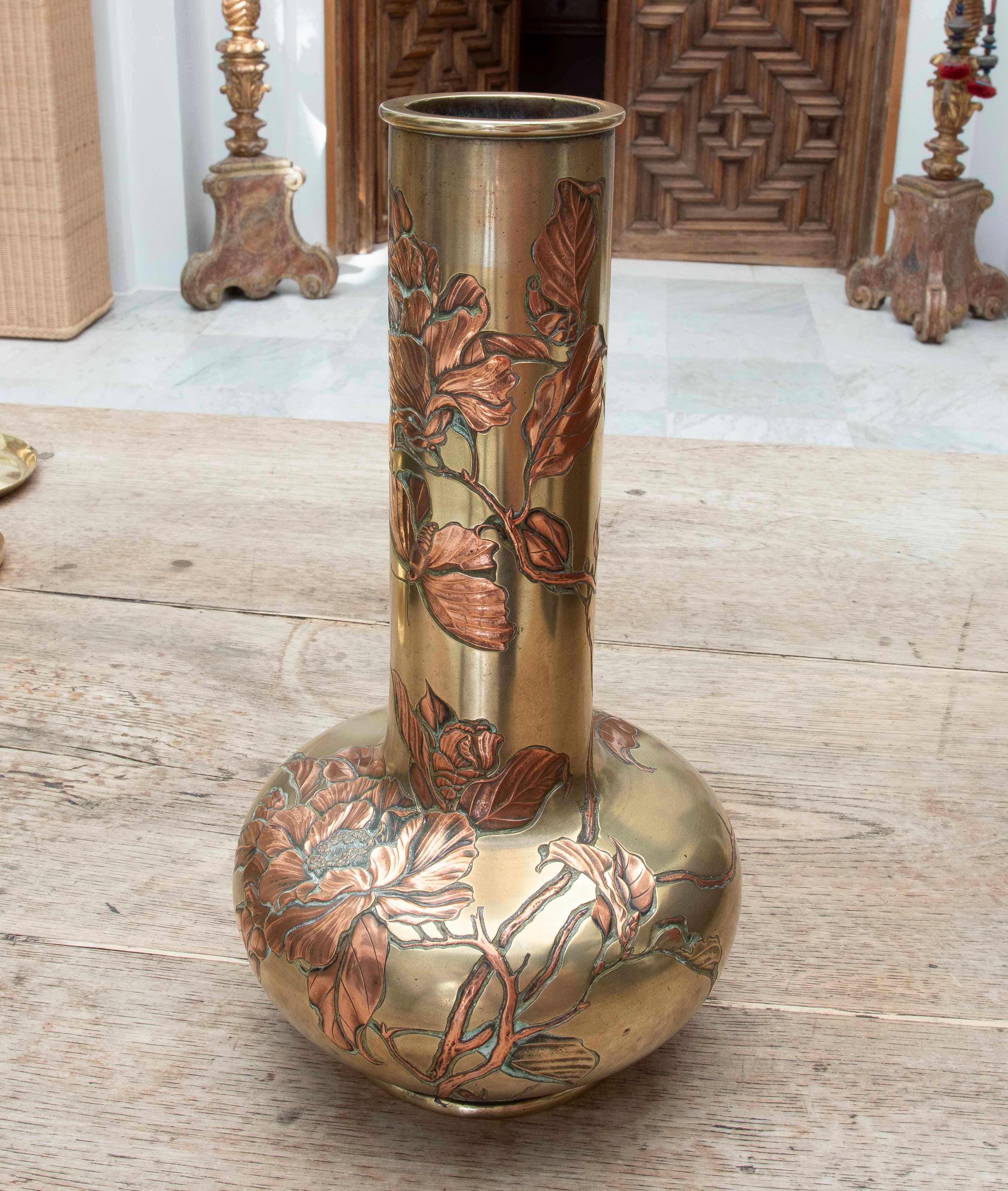 An elegant Japanese vase from the Meiji period, crafted in polished brass with embossed copper applications (repoussé), depicting a delicate plant motif with open flowers, sinuous leaves, and a butterfly in flight. The shape of the vase, with a