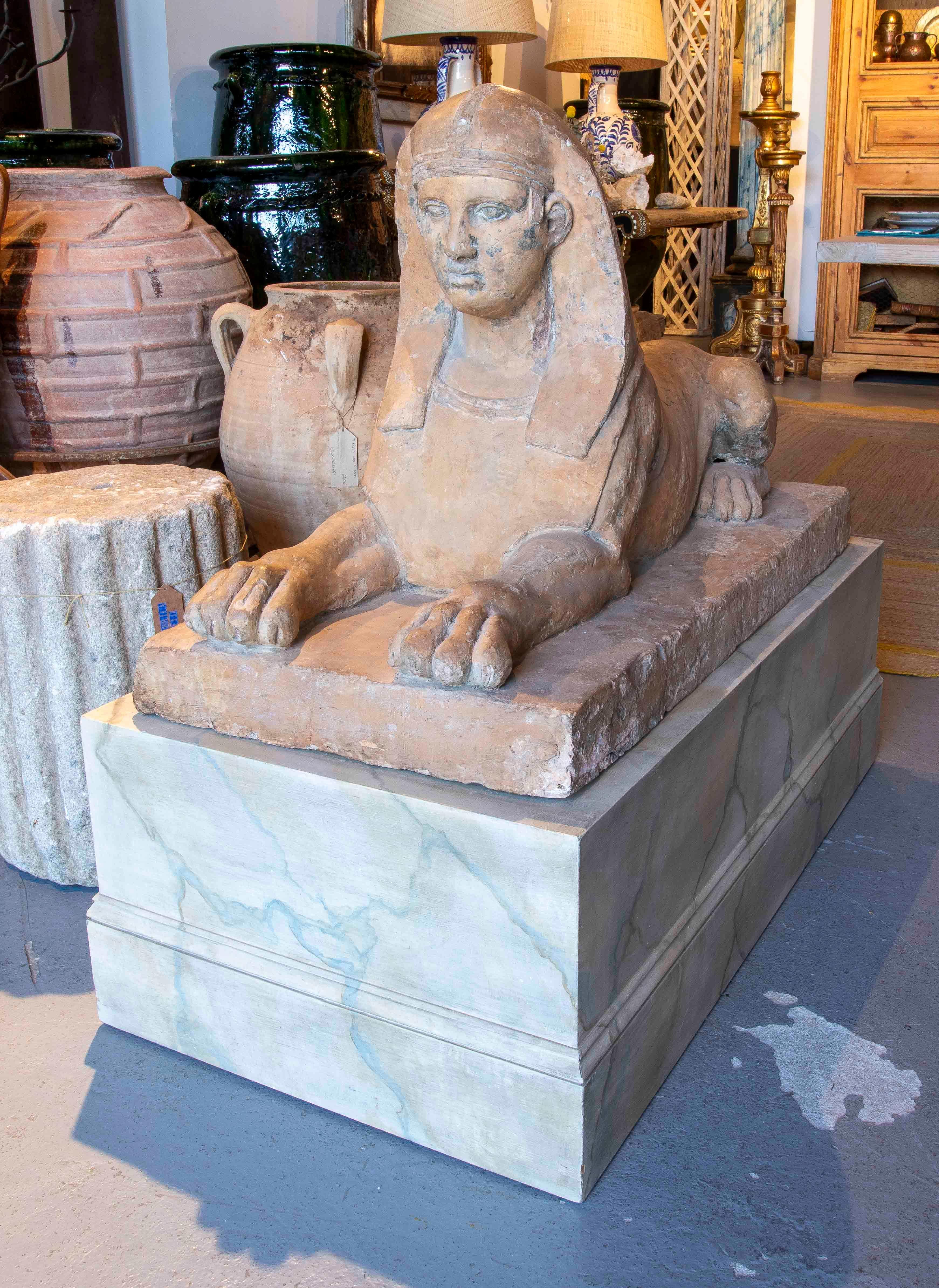 Magnifique sculpture de sphinx en terre cuite, représentée en position allongée avec une expression sereine et des détails bien définis sur le visage et les jambes. La pièce est montée sur un socle rectangulaire en bois marbré d'inspiration