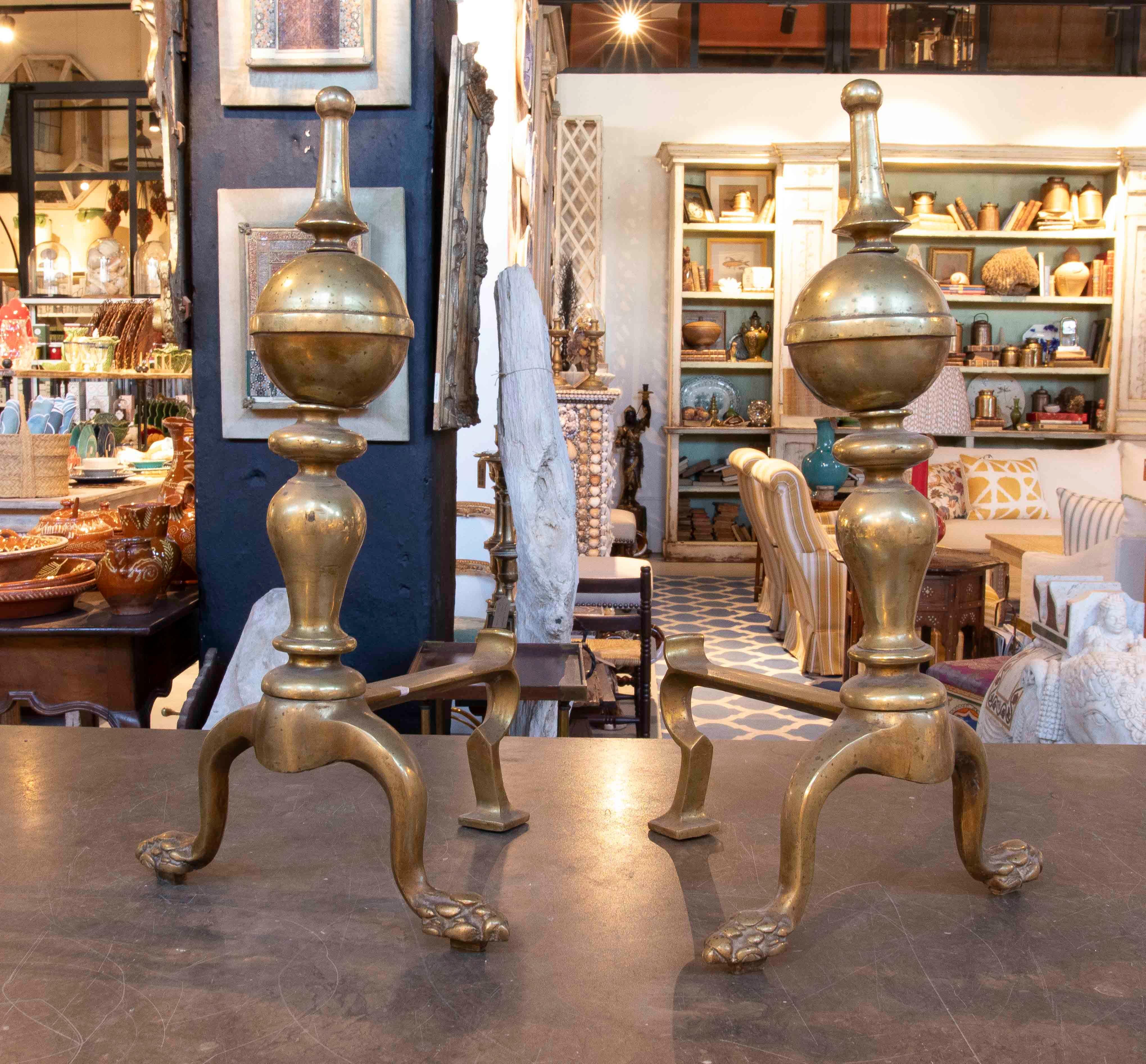 An elegant pair of antique andirons in solid brass, dating from the 19th century. Their classic design features upper spheres crowned with turned finials and legs shaped like feline claws, a symbol of strength and nobility. Their natural patina