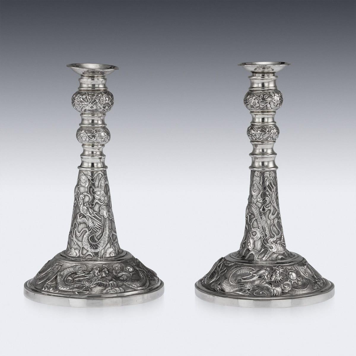 Paire de chandeliers décoratifs chinois en argent massif, de forme traditionnelle, avec un large pied et des becs amovibles, le sommet décoré de fleurs épanouies sur fond mat et la base de dragons entrelacés parmi des nuages, chassant la perle de la