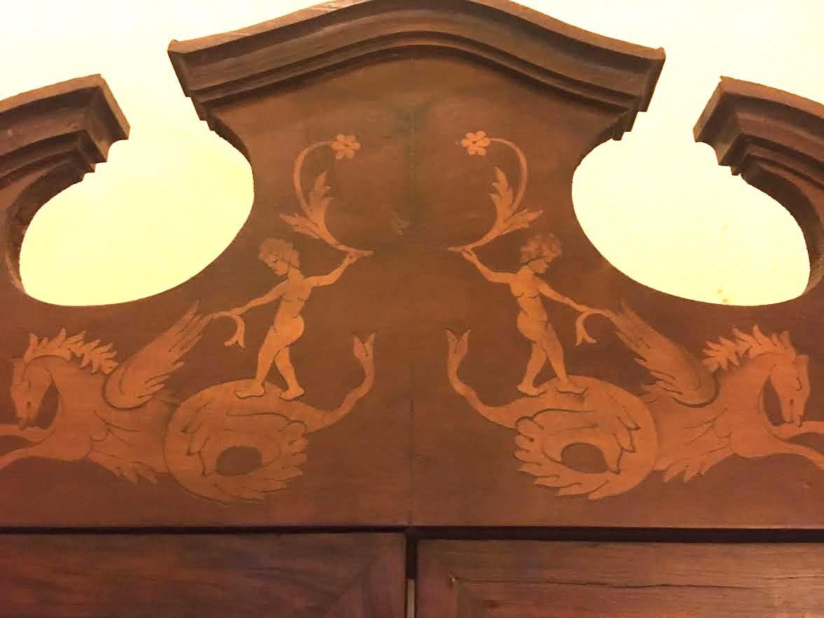Questa graziosa credenza angolare presenta ante in vetro su due ripiani ed è realizzata in noce con intarsi in legno di frutta. Fantasiosi putti a cavallo di ippocampi adornano il grazioso cornicione, un vaso di fiori decora l'anta inferiore con