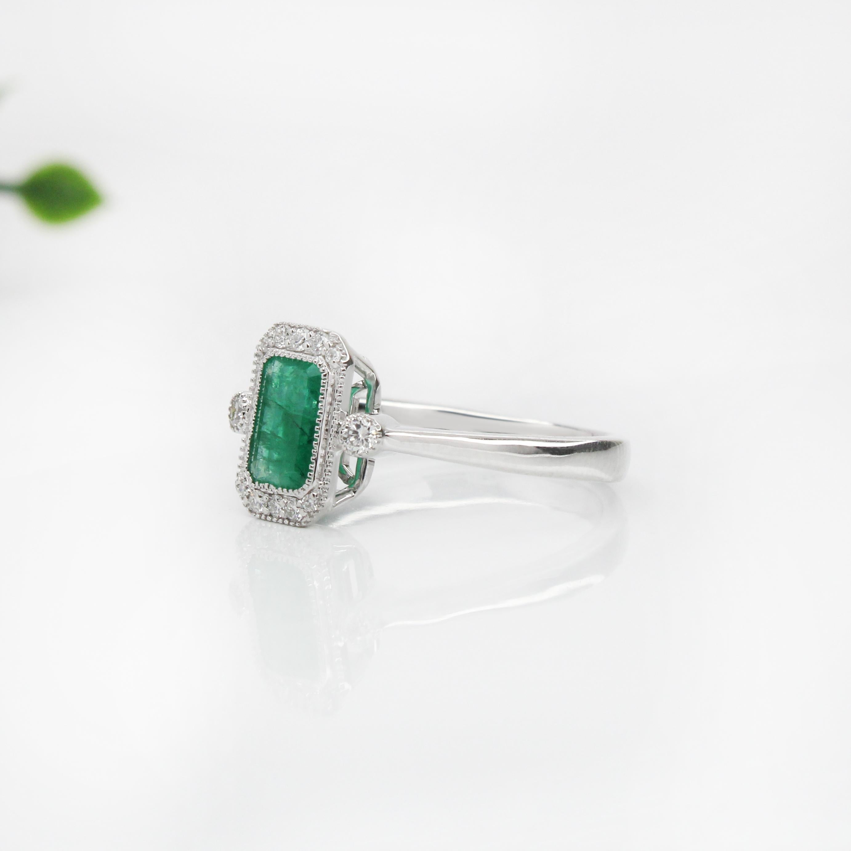 Dieser exquisite Ring präsentiert einen Smaragd von 1,2 Karat, der elegant von einem Kranz funkelnder Diamanten umrahmt ist. Der Smaragd ist in ein schlankes, poliertes Band eingefasst, das seinen leuchtend grünen Farbton hervorhebt. Das Design des