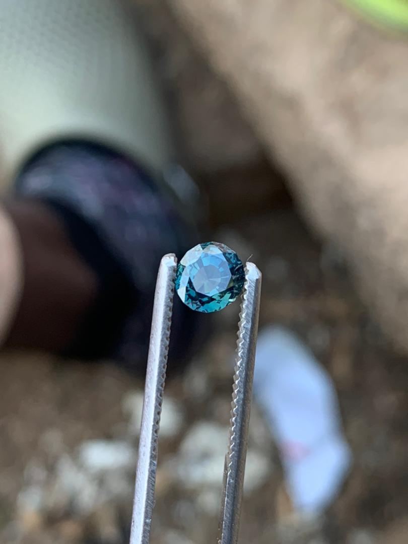 Discover the timeless beauty of this EYE CLEAN 1-carat Round Teal Blue Natural Untreated Sapphire Gemstone. Its classic round cut radiates with a symphony of light, enhancing the stone’s deep teal blue color that mirrors the serene hues of a