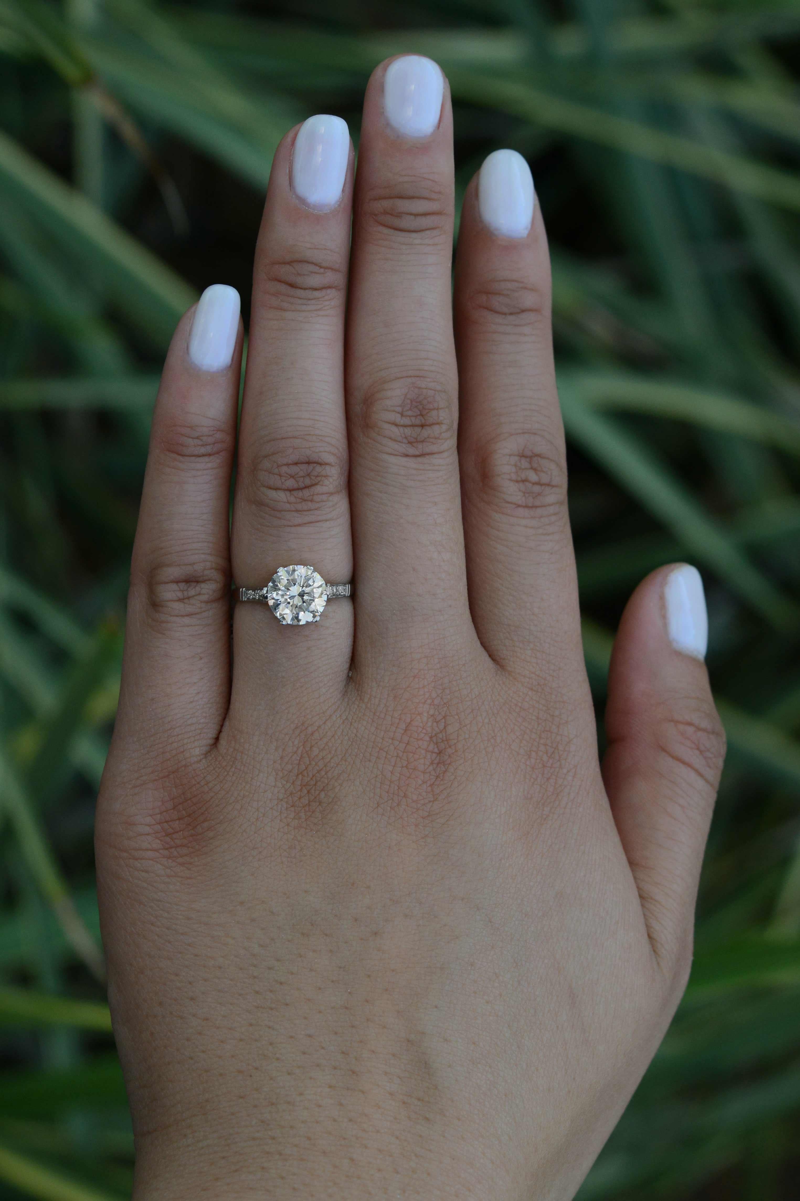 Una belleza Art Decó espectacular y sin esfuerzo. El alma de este anillo de compromiso vintage es un deslumbrante diamante brillante redondo casi incoloro de 2,5 quilates, dotado de un brillo y un encanto increíbles. El encantador engaste de platino