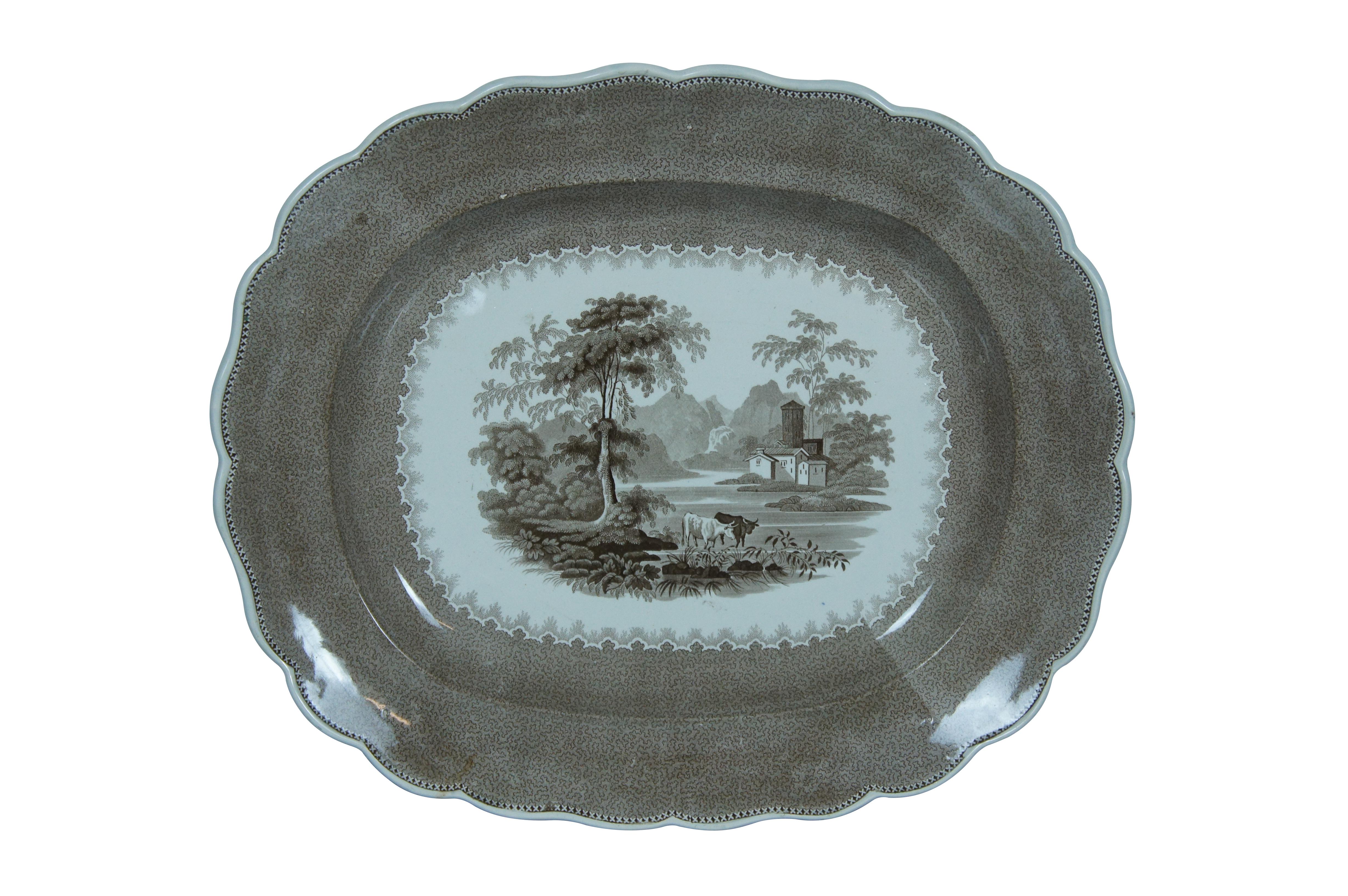 Set of two late 19th century brown transferware serving platters. One scalloped edge, rounded rectangular platter of unknown maker, marked Scroll B, showing an idyllic landscape of cows in a mountain lake surrounded by a patterned border. Oval