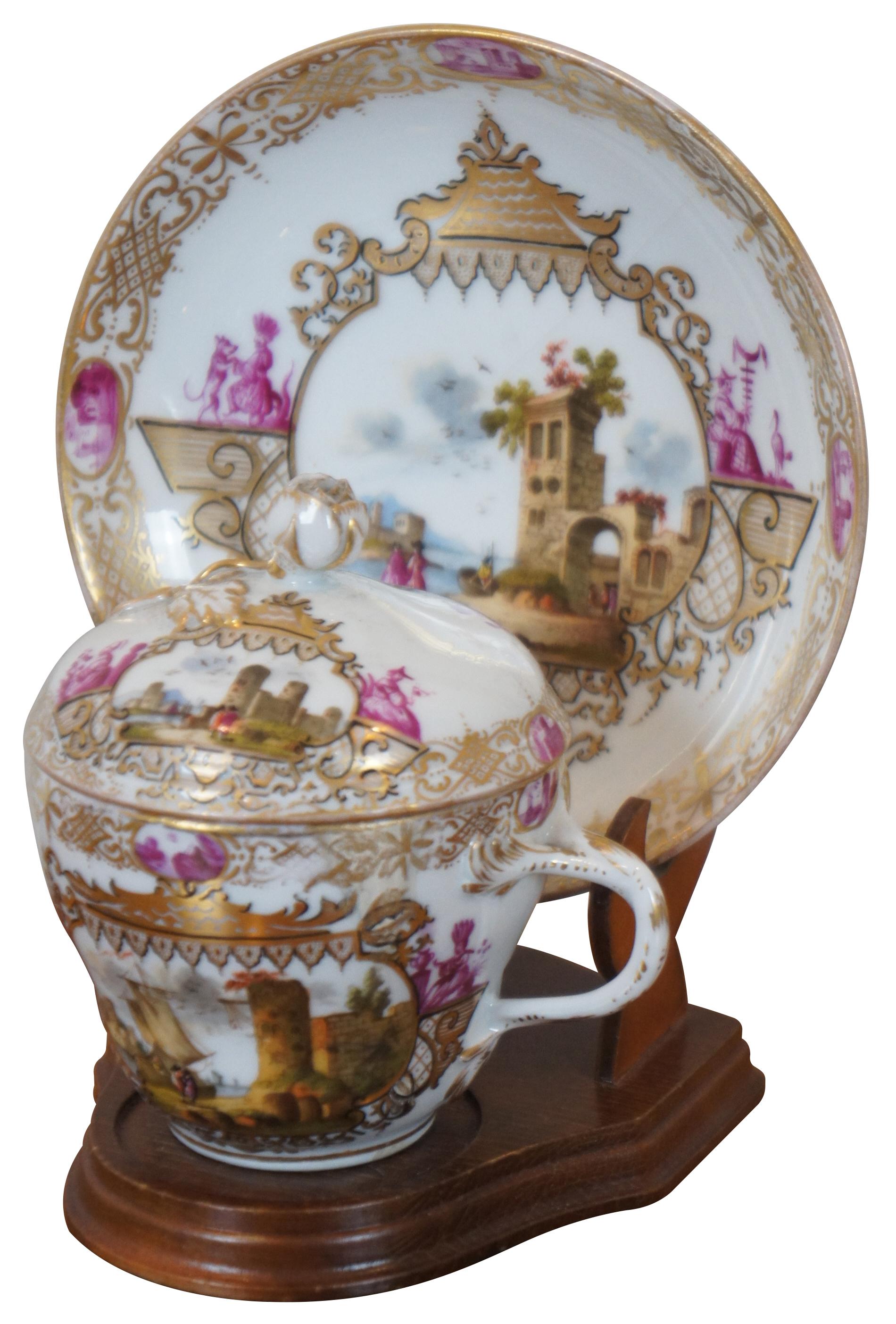 Pair of exquisite antique 19th century Meissen porcelain demitasse chocolate cups with lids and saucers, decorated with pink and gold borders and landscapes with stone buildings. Both are beautifully displayed on wooden stands.

