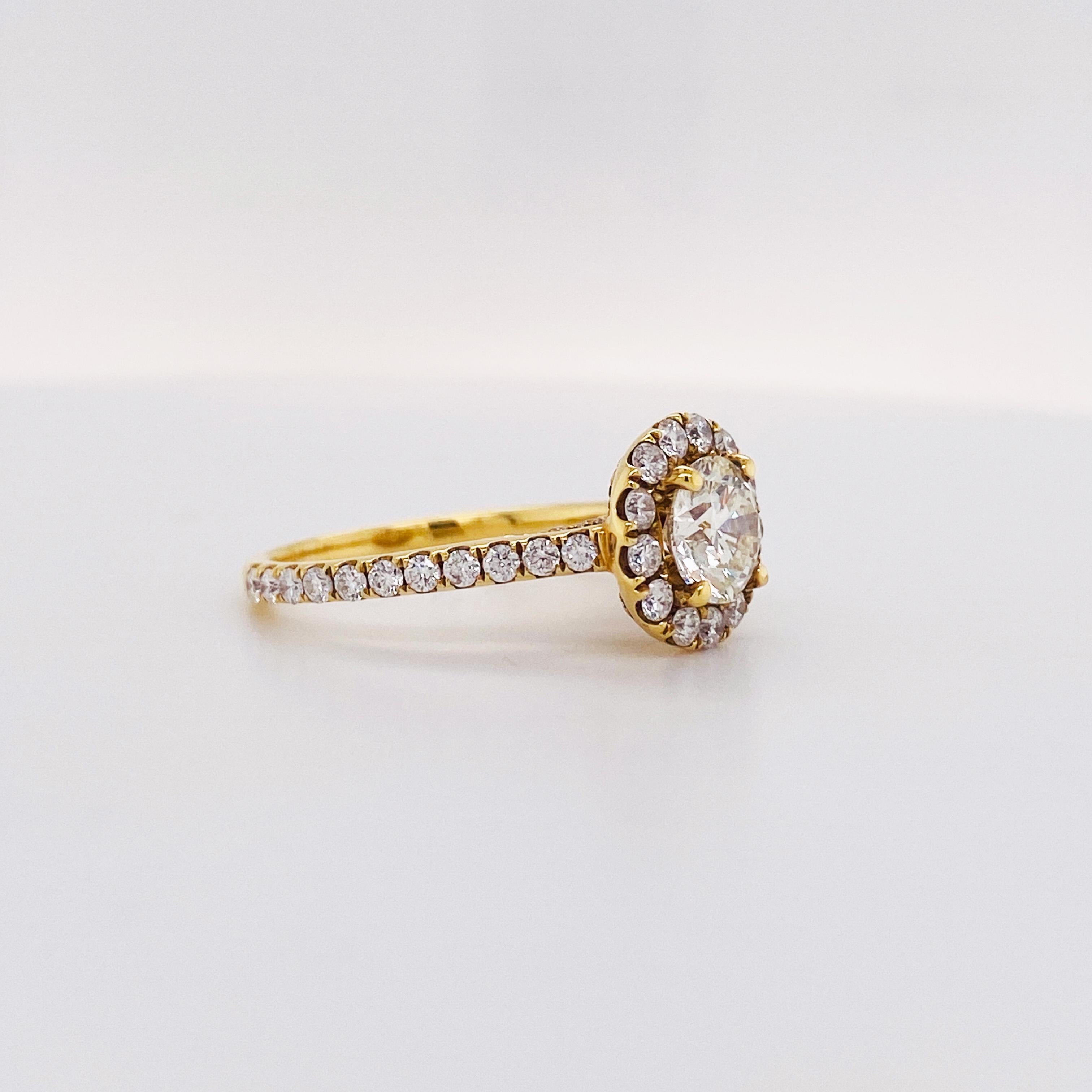 This 18 karat yellow gold diamond engagement ring holds a 1.00 carat round brilliant diamond, SI2 clarity and H color with a diamond halo surrounding the center stone. The ring has a diamond band that goes 3/4 around the ring and comes up in a