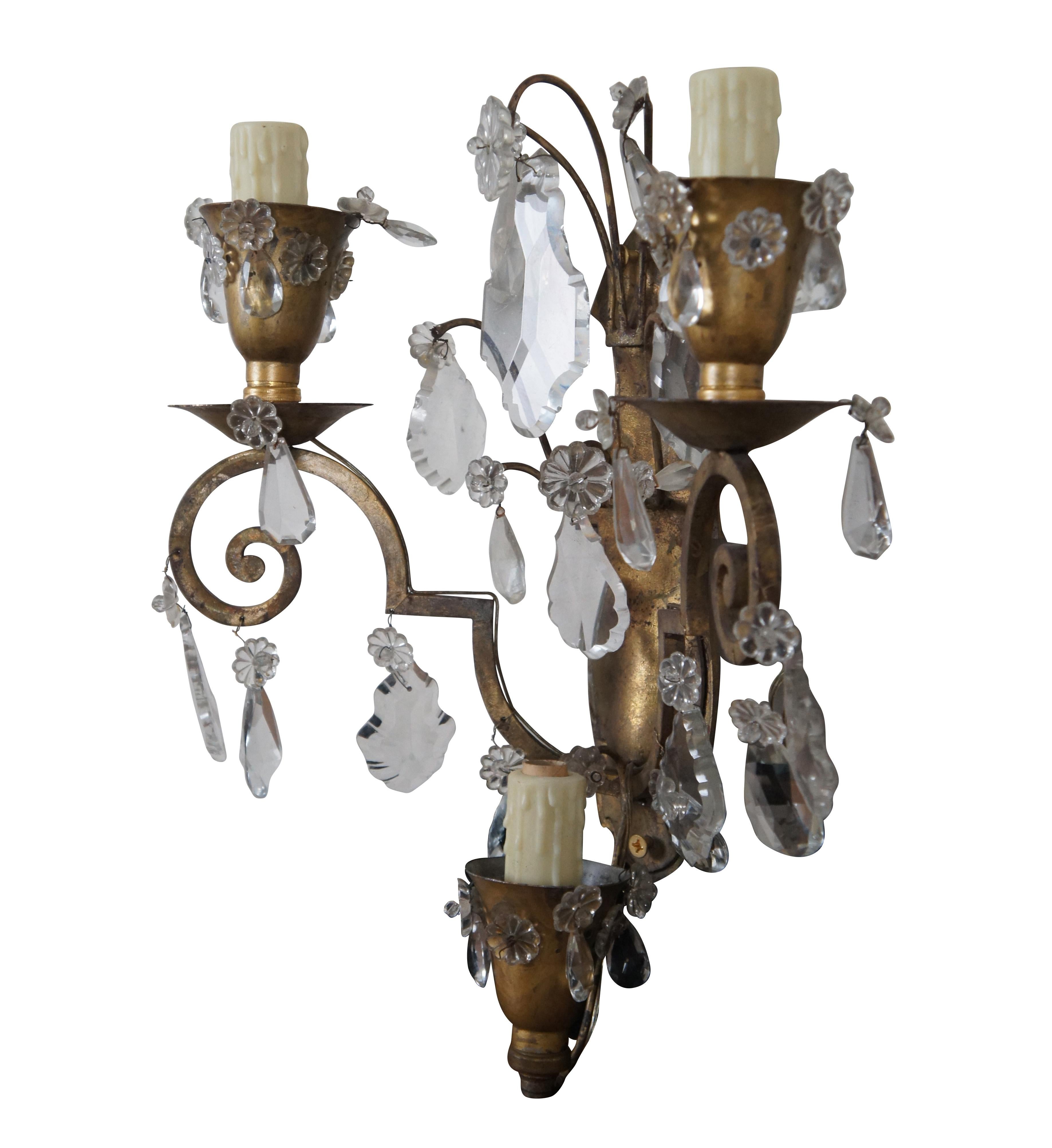 A stately pair of Hollywood Regency gilded brass and drop crystal three light wall sconces. Features scrolled arms, realistic candle covers, Swedish drop crystals, pendeloques, and floral crystal medallions accents. 2 sets available.