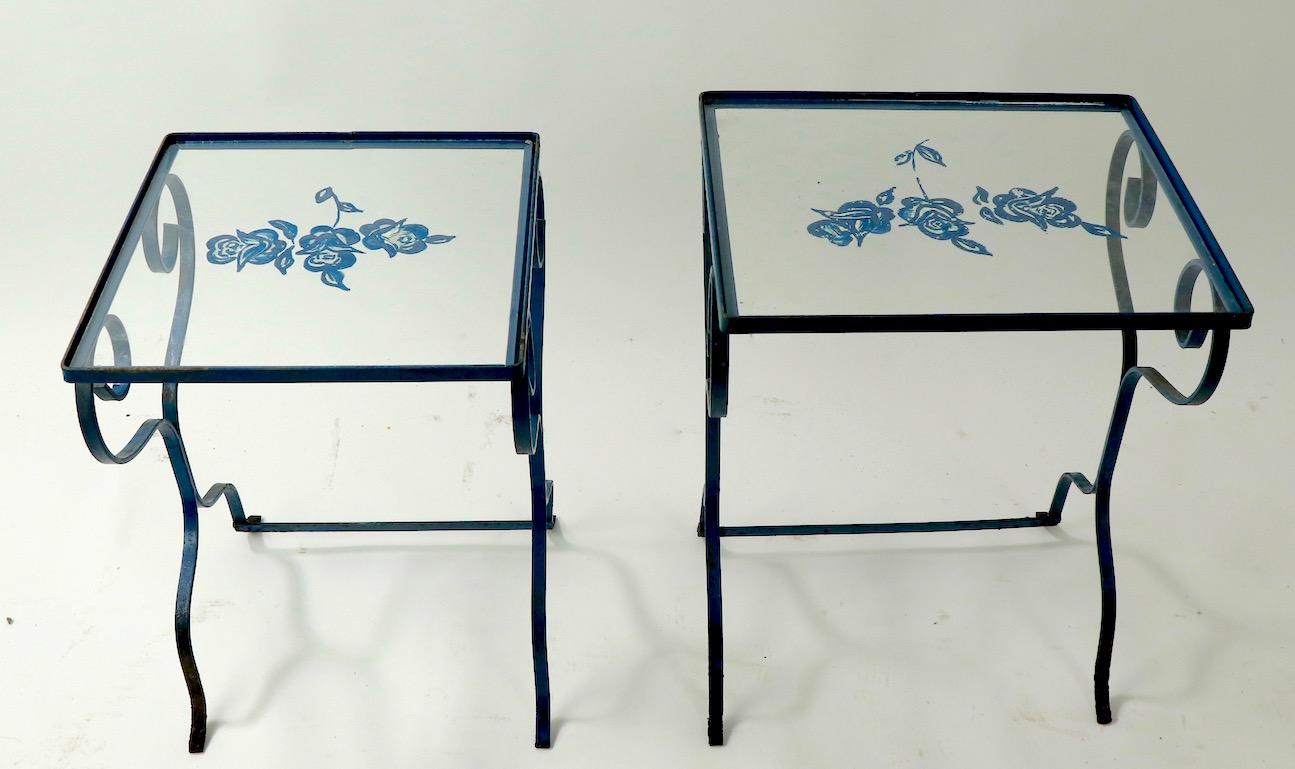 Set of 2 wrought iron nesting tables attributed to Salterini, each having glass tops with hand painted floral decoration. The tables are in good, original condition, the feet show surface rust, and the floral paintings have minor loss, as shown.