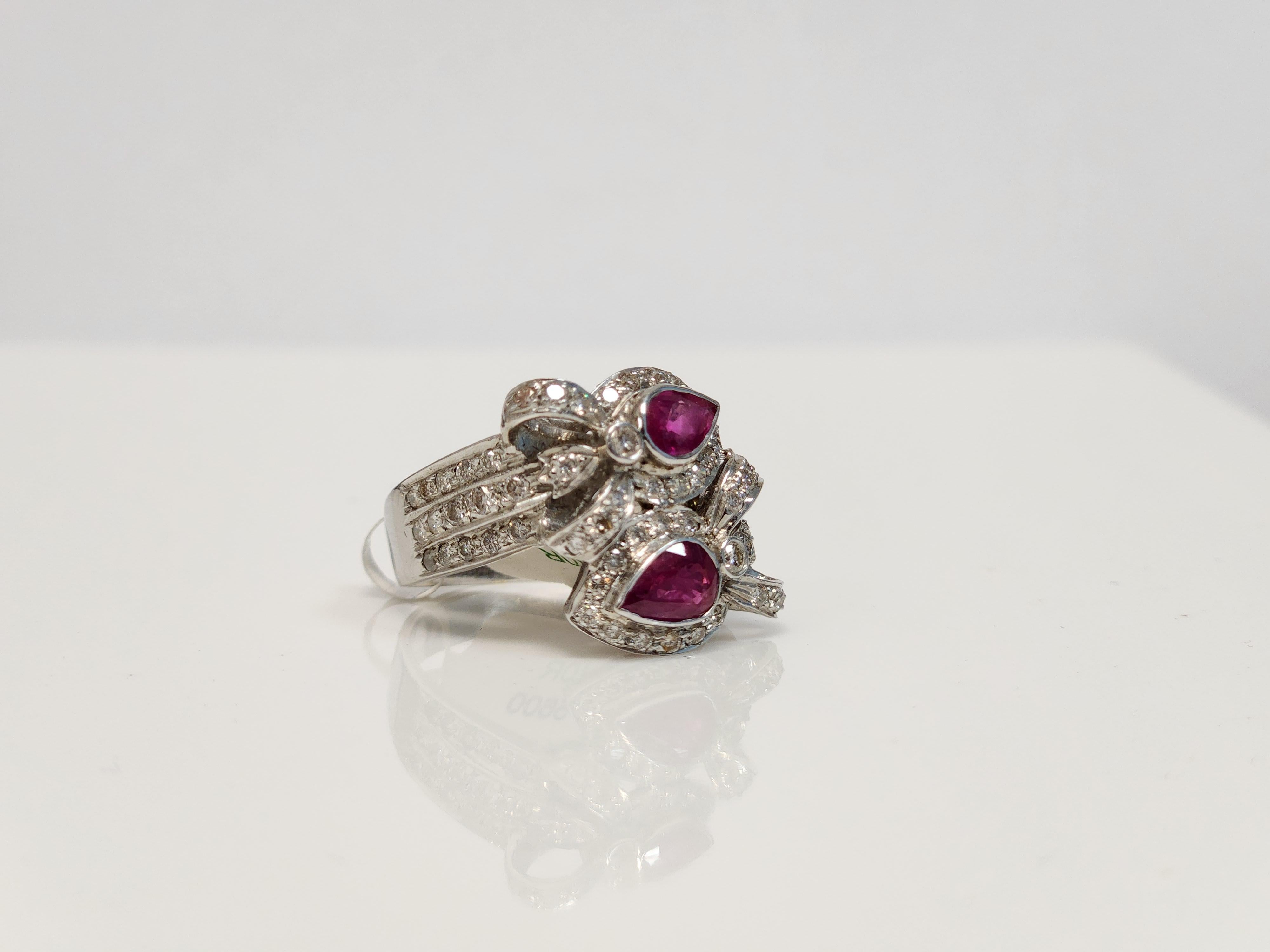 Indulge in the allure of vibrant red with this exquisite 2 Pear Shaped Red Ruby Bypass Ring. The two 0.65 carat pear-shaped red rubies elegantly bypass each other, surrounded by a halo of 1.05 carat white melee diamonds that add a stunning sparkle