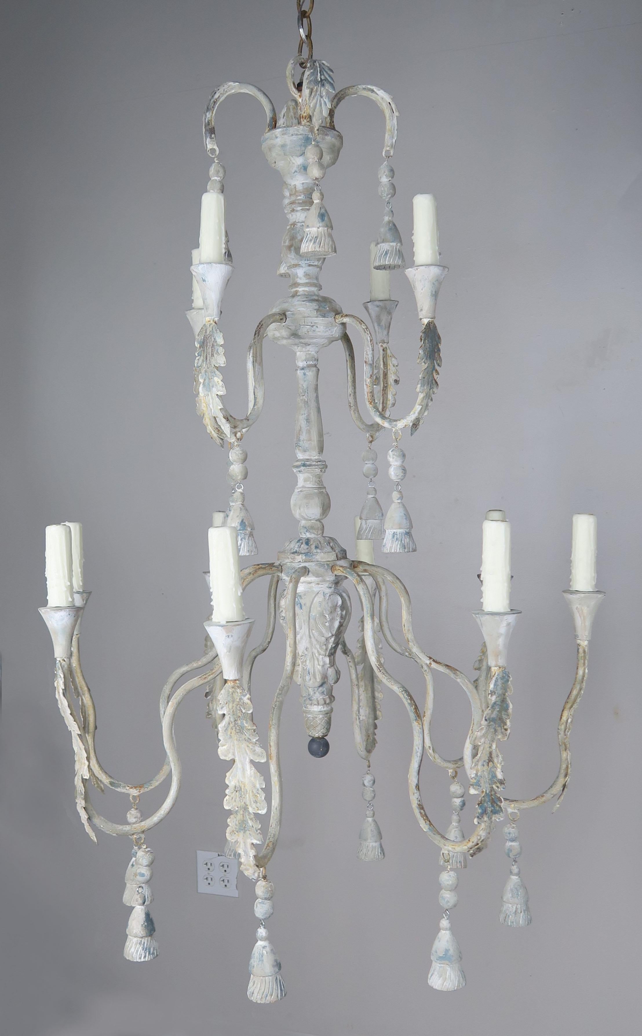 Pair of Italian two-tier twelve-light Italian style painted chandeliers with carved wood tassels and beads throughout. The paint on the fixtures is beautifully worn with an antique colored cream base and remnants of soft blue paint, rust and natural