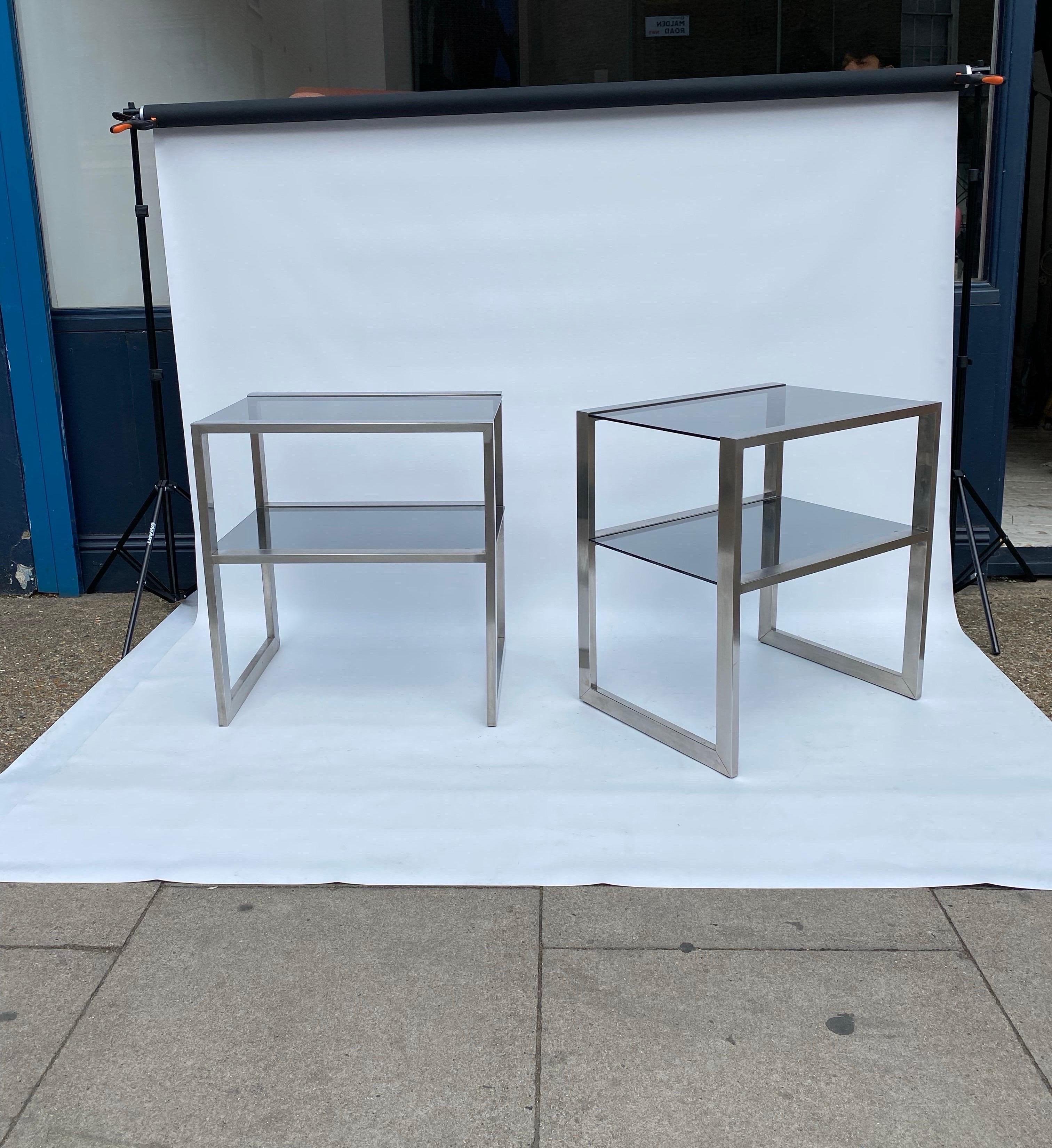Mesas auxiliares de dos niveles con estructura de metal y estantes de cristal ahumado gris, atribuidas a Guy Lefevre para Maison Jansen en la década de 1970. Tiene una estética minimalista, con un marco de acero cepillado y encimeras de cristal