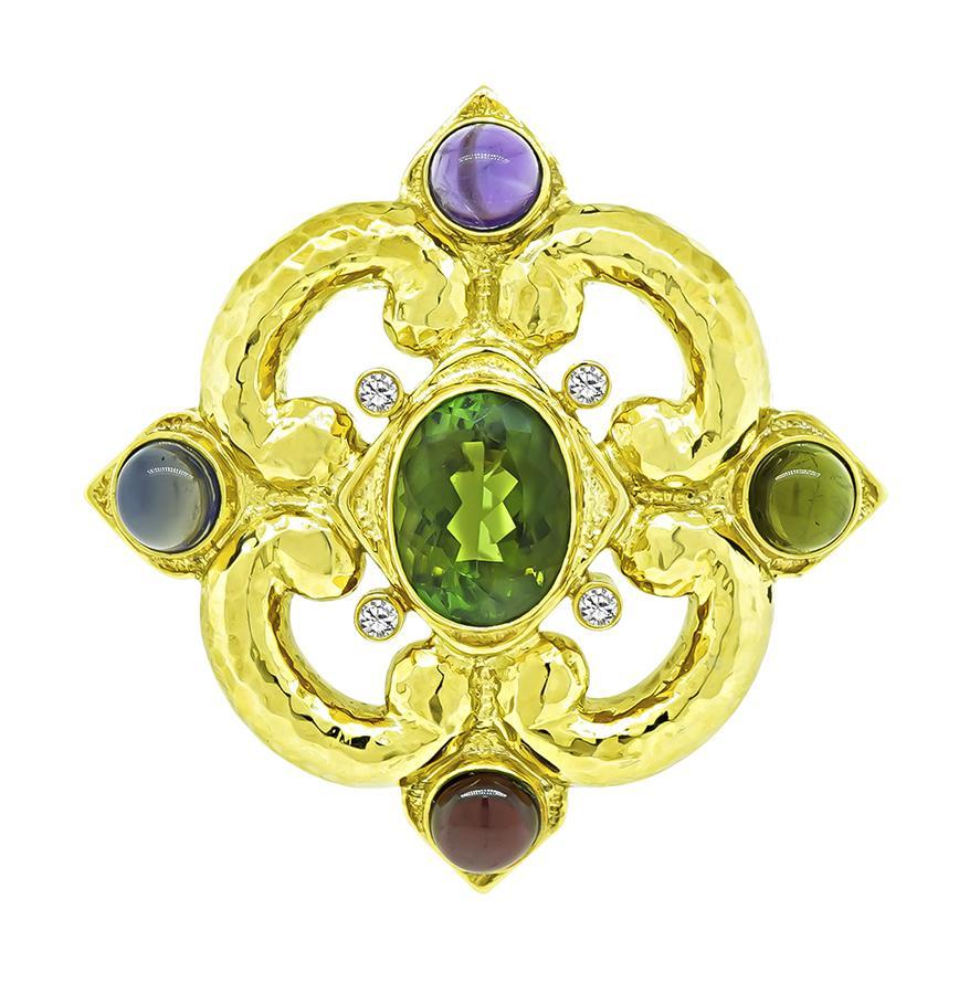 Si tratta di una splendida spilla/pendente in oro giallo 18 carati. La spilla è centrata da una bella tormalina verde di taglio ovale che pesa circa 20,00ct. La pietra centrale è accentuata da iolite cabochon, ametista, peridoto, granato e diamanti