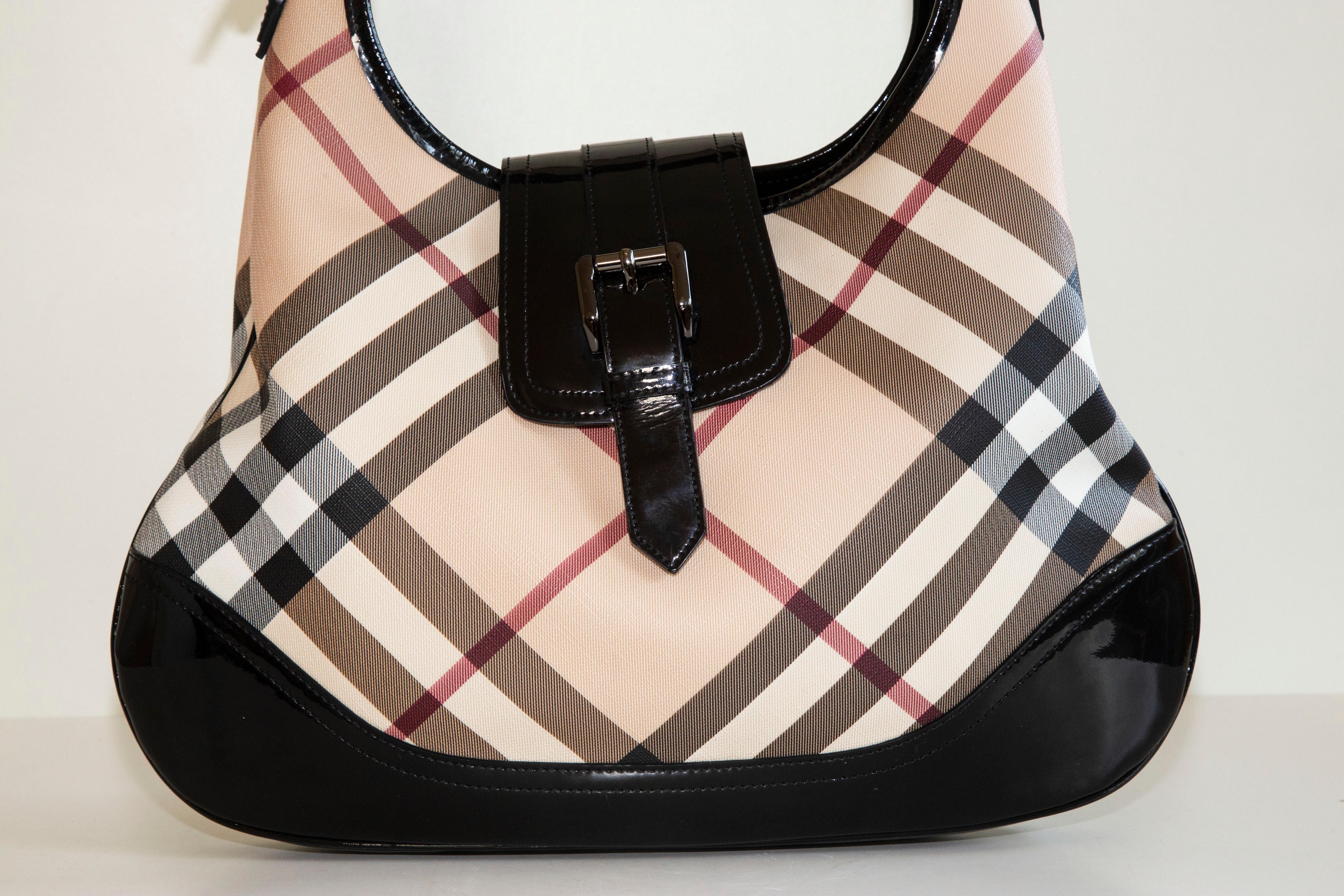 An authentic Burberry Brook hobo shoulder bag. The bag features a Nova Check coated canvas, black patent leather trim, and pewter-toned hardware. The interior is lined with beige fabric and next to the major compartment,  there are three side