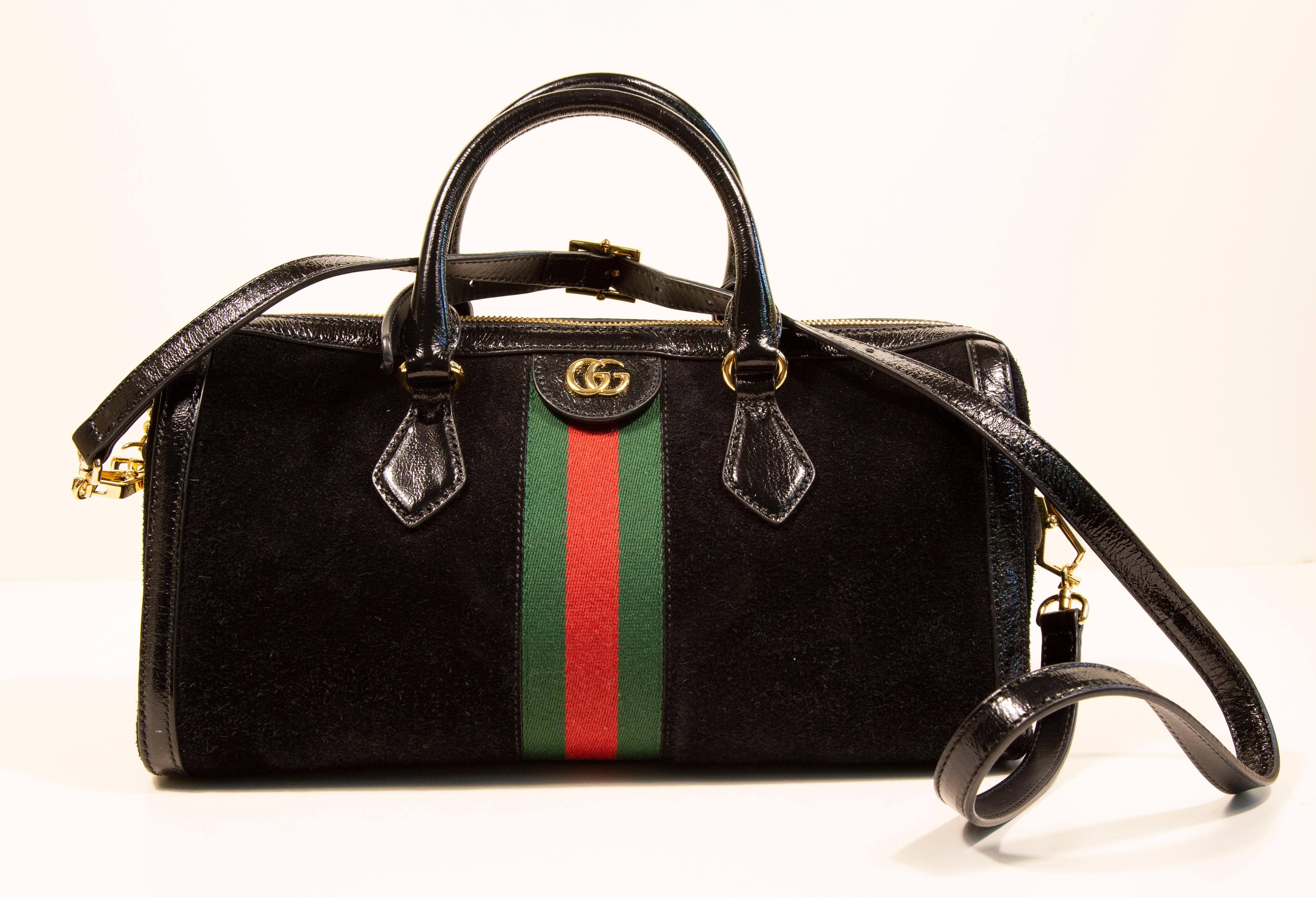 Gucci Ophidia Boston bag. The bag is made of black suede and black patent leather trim with gold toned hardware. It features the Gucci signature race strap at the front and the back side of the bag. The interior is lined with beige suede like
