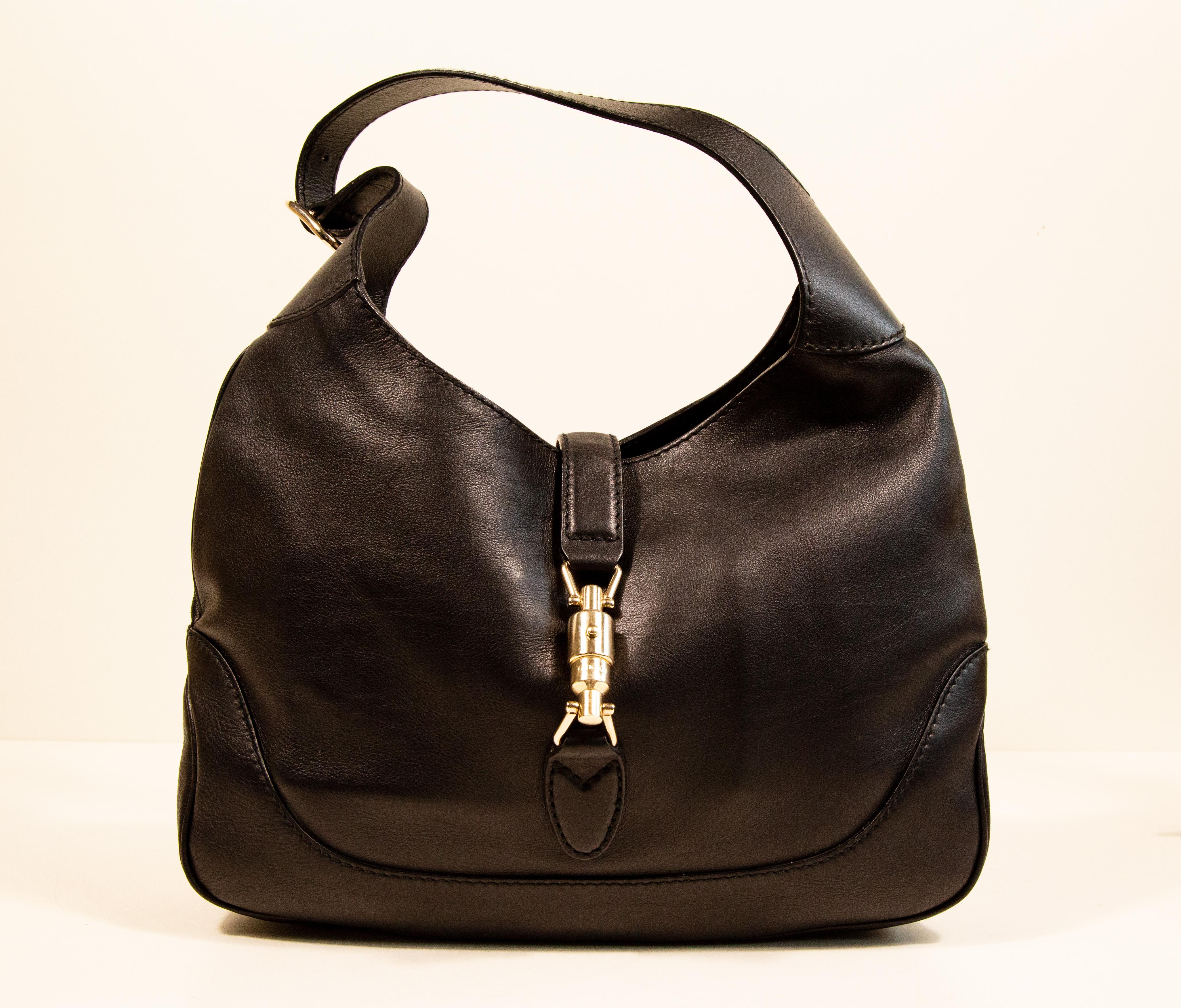 Una Jackie Gucci de piel negra con herrajes en tono dorado claro. El interior está forrado de tela beige, y junto al compartimento principal presenta tres bolsillos laterales, uno de ellos con cremallera. La bolsa puede cerrarse con el cierre de