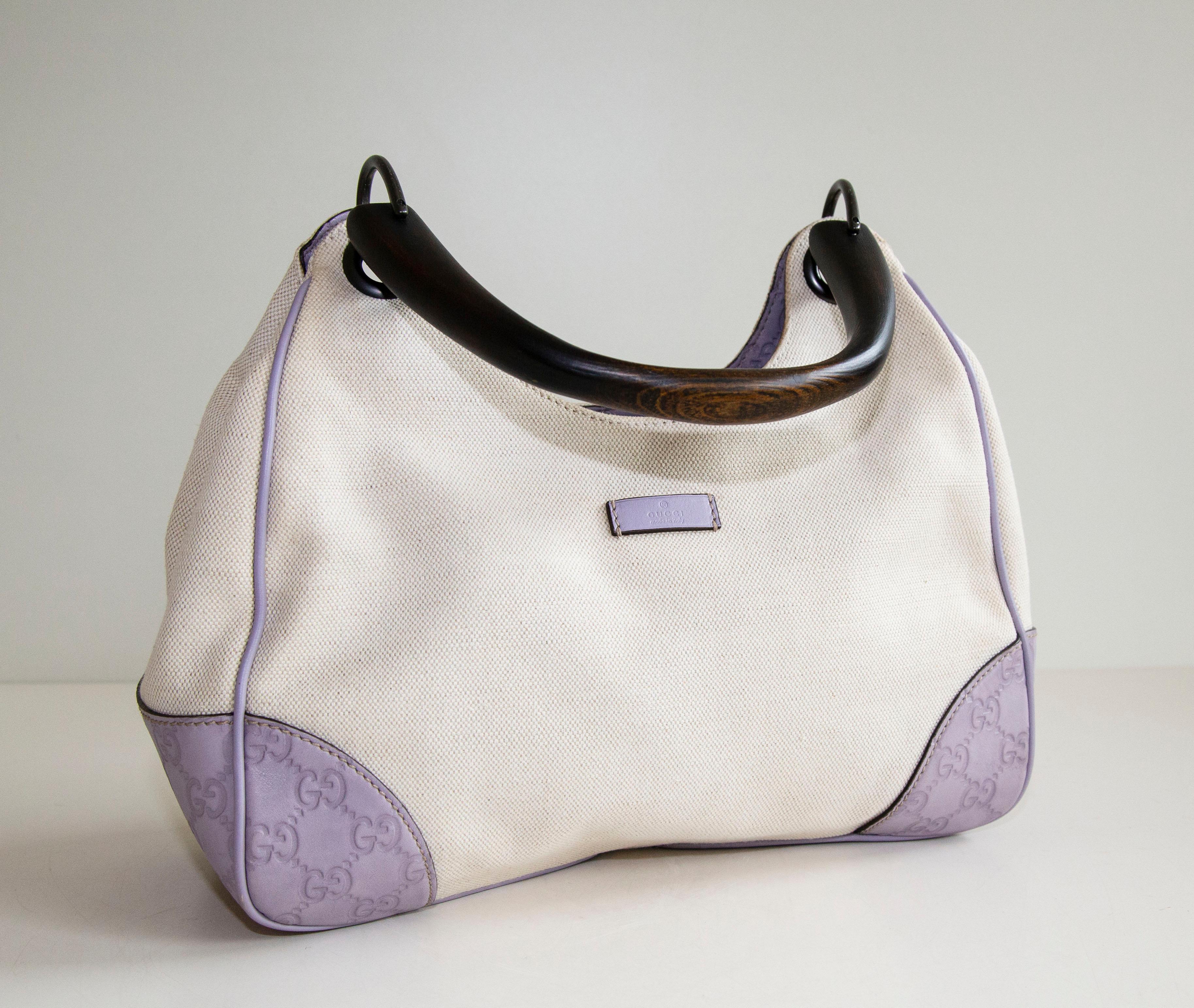 An authentic Gucci Shima wood top handle bag. The bag features a purple Guccisima trim, a beige canvas exterior, and a wooden handle. The interior is lined with black fabric and there is one side pocket with a zipper. The interior is neat & clean.
