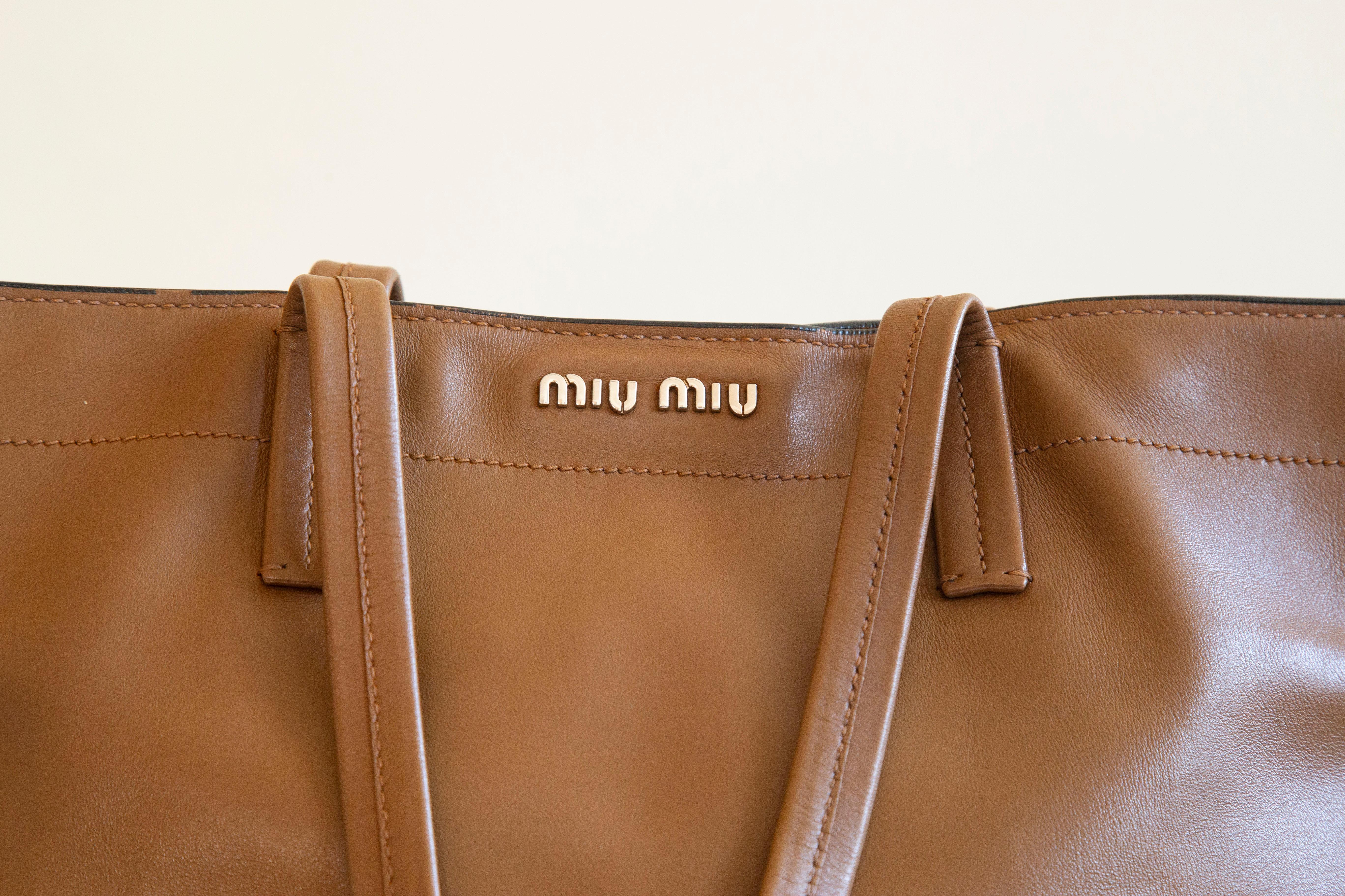 An authentic Miu Miu tote bag. The bag features a light brown leather exterior and silver-tone hardware. The interior is lined with black fabric and there are three side pockets of which one has a zipper. There is also an additional zipped pocket on