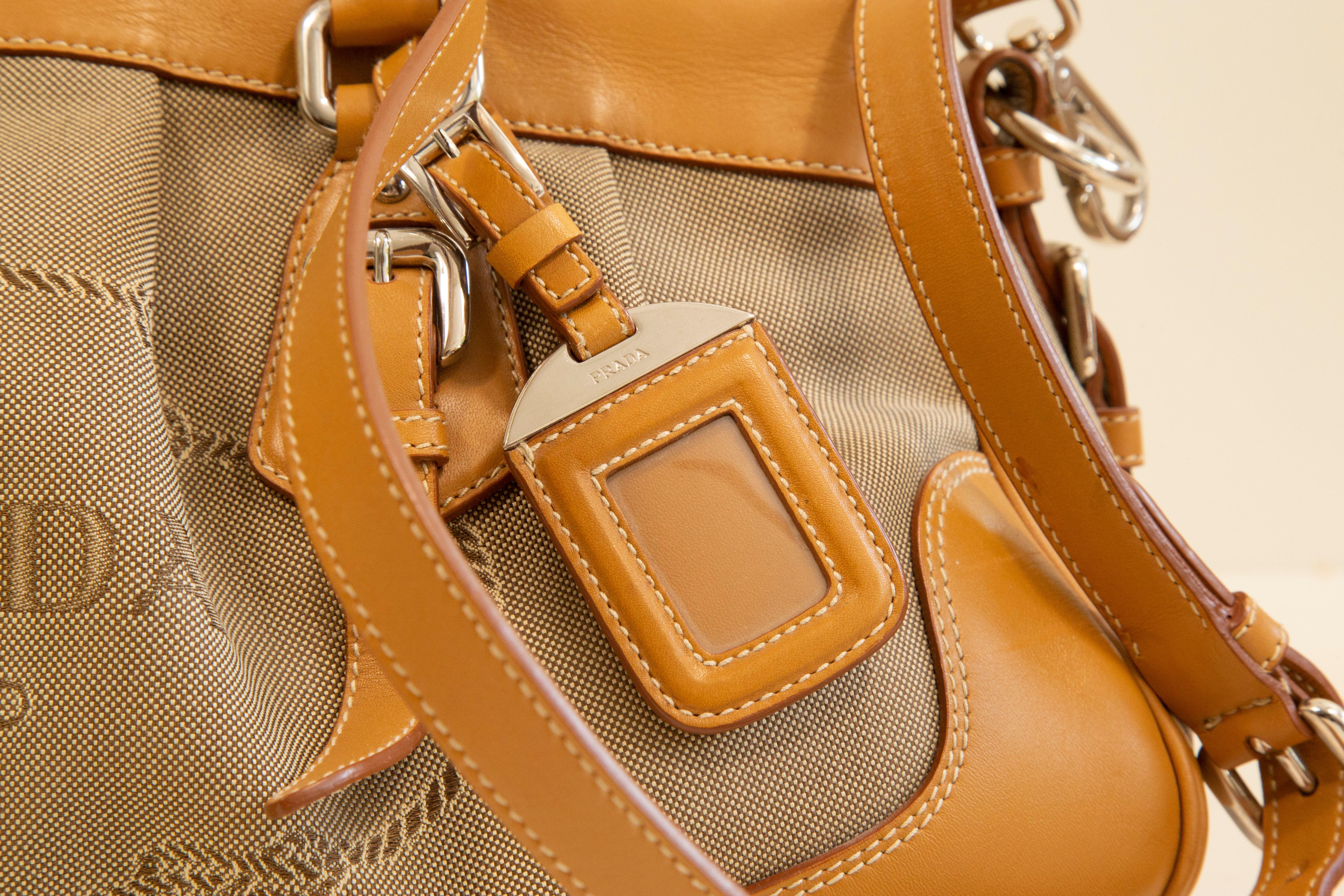 Prada 2-way bag that can be worn as a top handle bag or as a shoulder bag/crossbody. The bag is made of light brown canvas with tan colored leather trim and silver toned hardware. The interior is lined with a synthetic fabric with Prada logo and