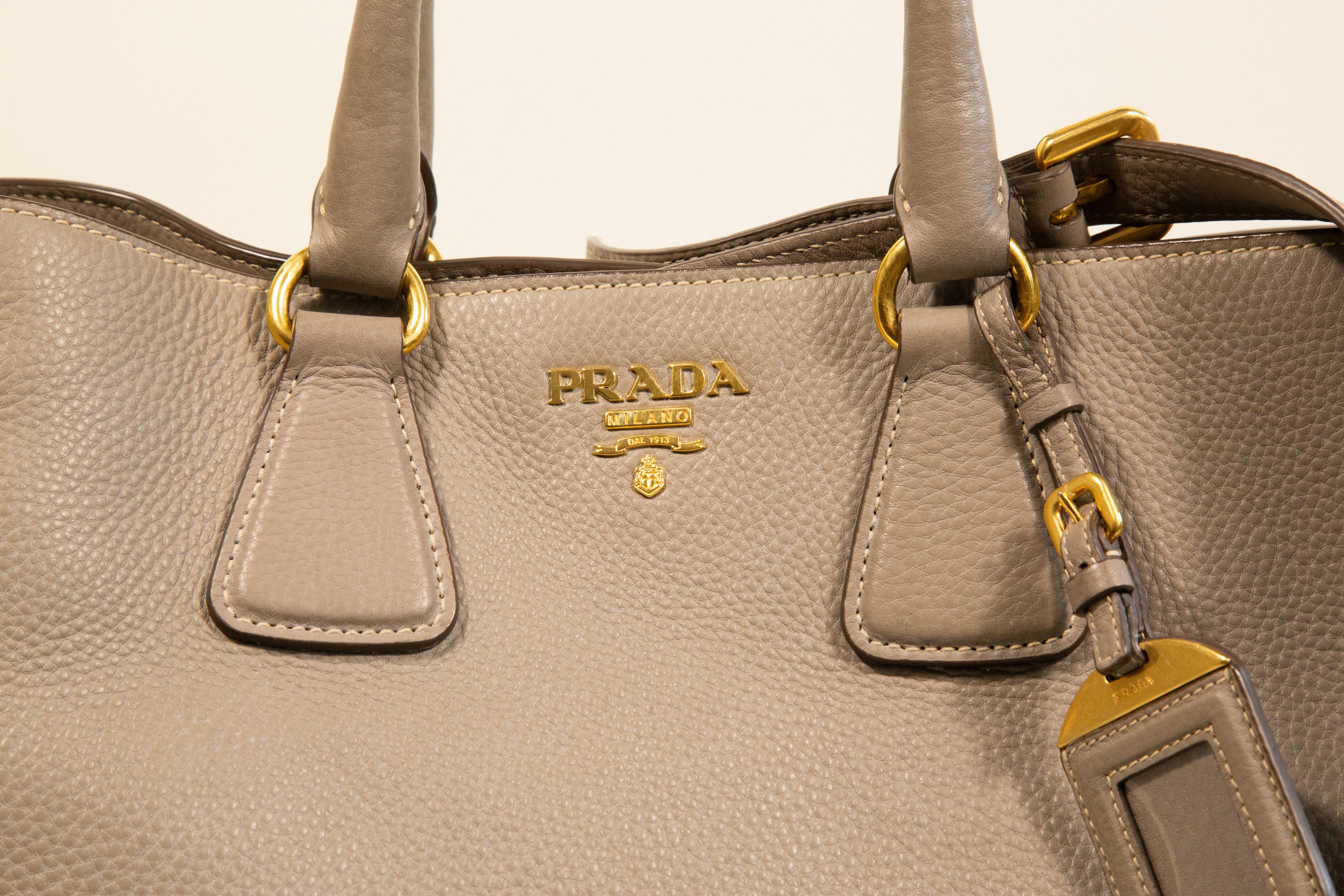 Prada 2-way bag made of gray leather with gold toned hardware. The bag is lined with brown Prada fabric and the interior consists of one major compartment with two side pockets of which one has a zipper. The bag can be worn as a top handle bag with