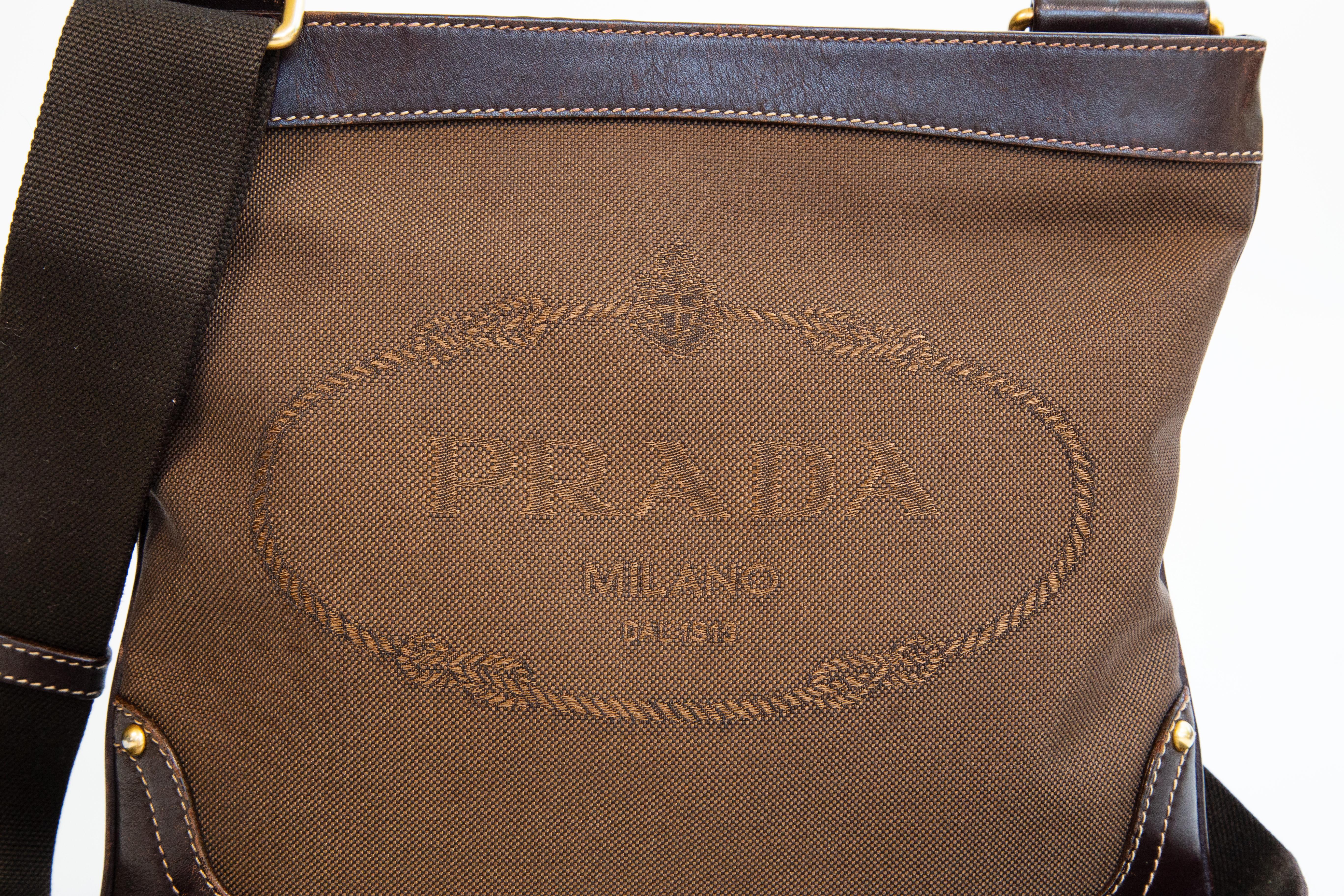 Sac à bandoulière Prada en toile marron et garniture en cuir marron avec des ferrures dorées. L'intérieur est doublé d'une doublure synthétique marron avec le logo Prada. A côté du compartiment principal, il y a une poche latérale avec une fermeture