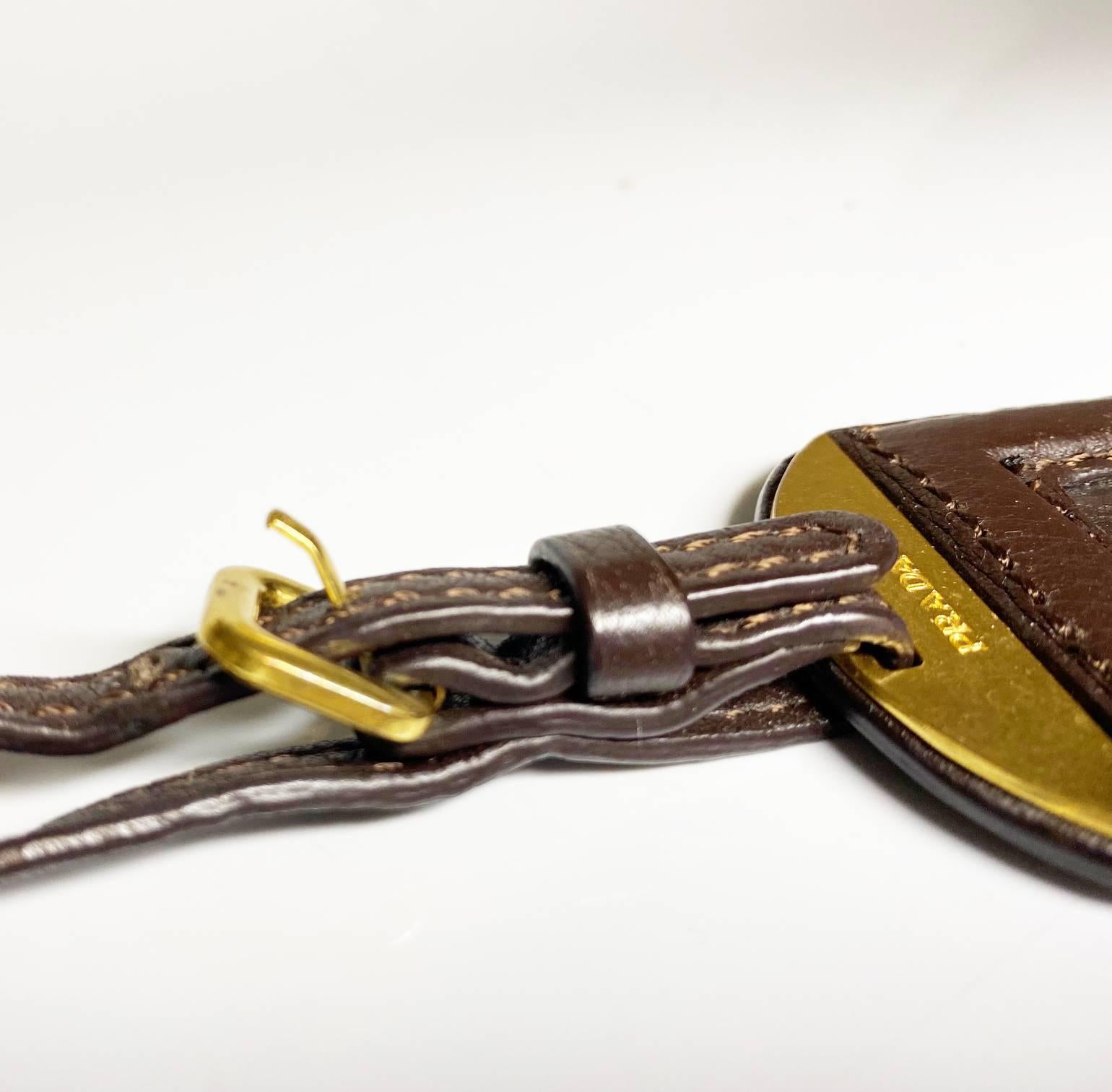  2000s Prada Chocolate Brown Leather Travel Tag adorned with opulent gold hardware and crafted from premium leather. With an adjustable strap length, this exquisite accessory exudes effortless elegance and undeniable