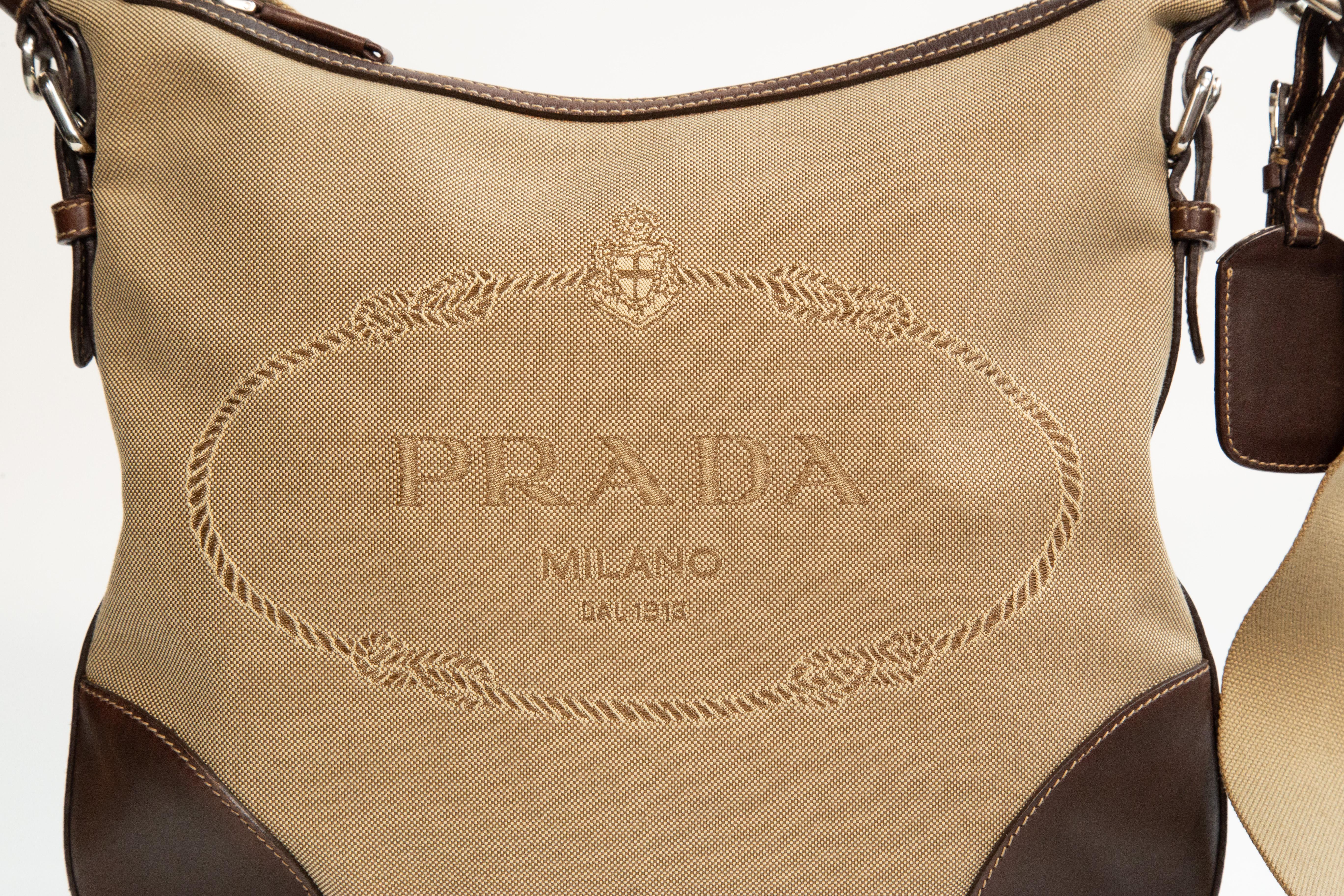 Borsa crossbody di Prada in tela con finiture in pelle marrone e finiture in argento. L'interno è foderato in tessuto sintetico marrone con logo Prada. Accanto allo scomparto principale, l'interno presenta una tasca laterale con cerniera. La borsa